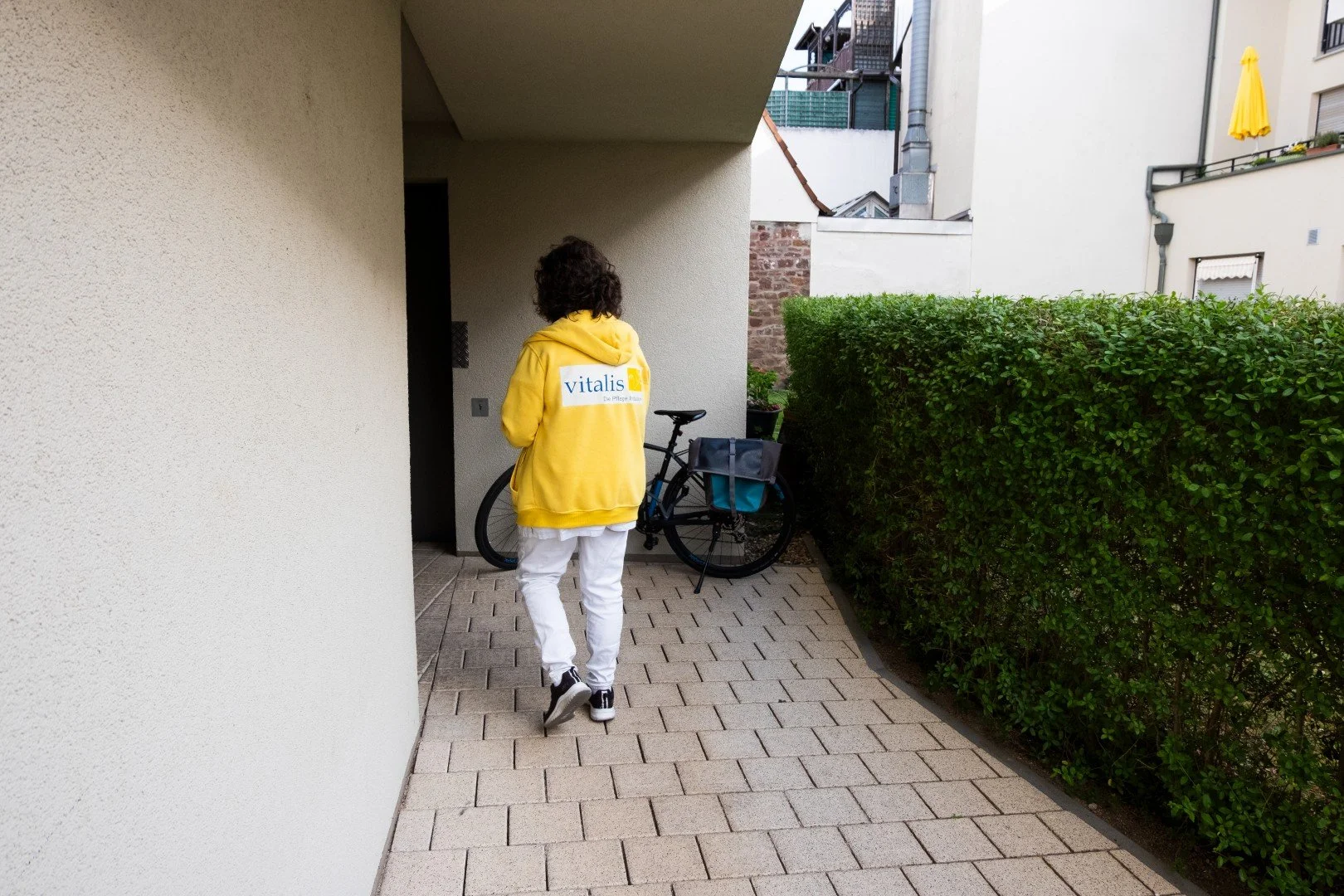 Eine Person in gelber Jacke mit dem Logo 'vitalis' beobachtet einen Fahrradständer vor einem Eingang eines Mehrfamilienhauses, umgeben von einer Hecke und anderen Gebäuden.