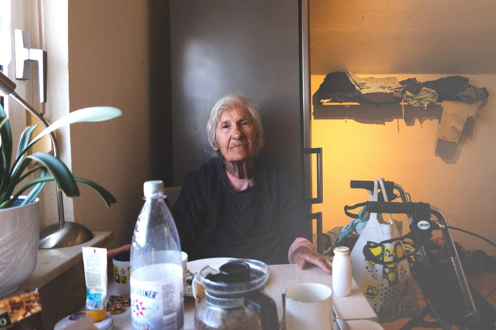 Ältere Frau sitzt an einem Tisch mit Getränken, vor ihr stehen Trinkflasche, Kaffeekanne und Tasse. Hinter ihr eine braune Tür, rechts eine Nische mit Kleidung und einem Rollator.