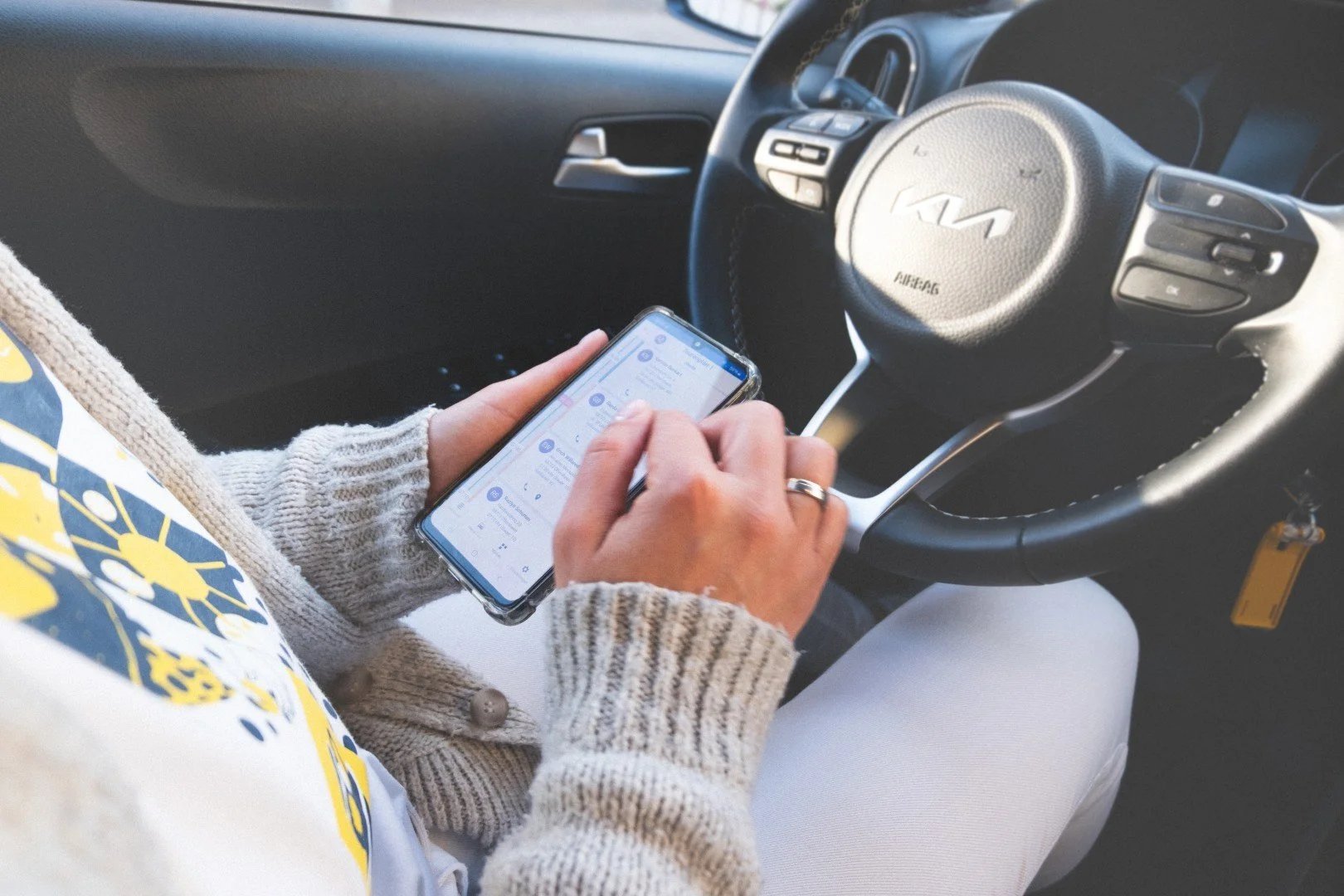 Person benutzt ein Smartphone im Auto, das Lenkrad einer Kia-Fahrzeug. Die Person trägt einen grauen Pullover und sitzt auf dem Fahrersitz.
