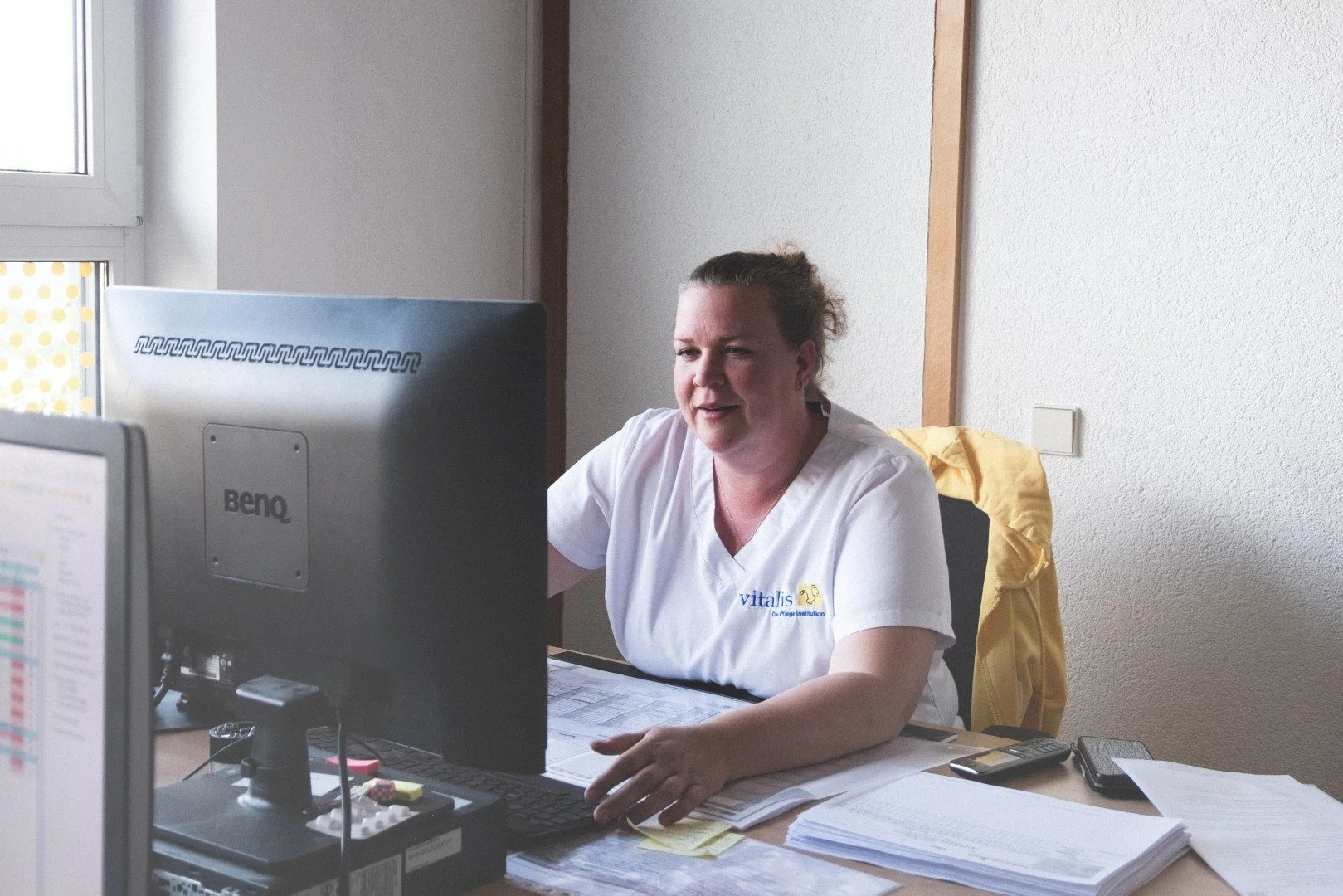 Eine Frau sitzt an einem Schreibtisch in einem Büro, arbeitet an einem Computer und trägt ein weißes medizinisches Uniform. 