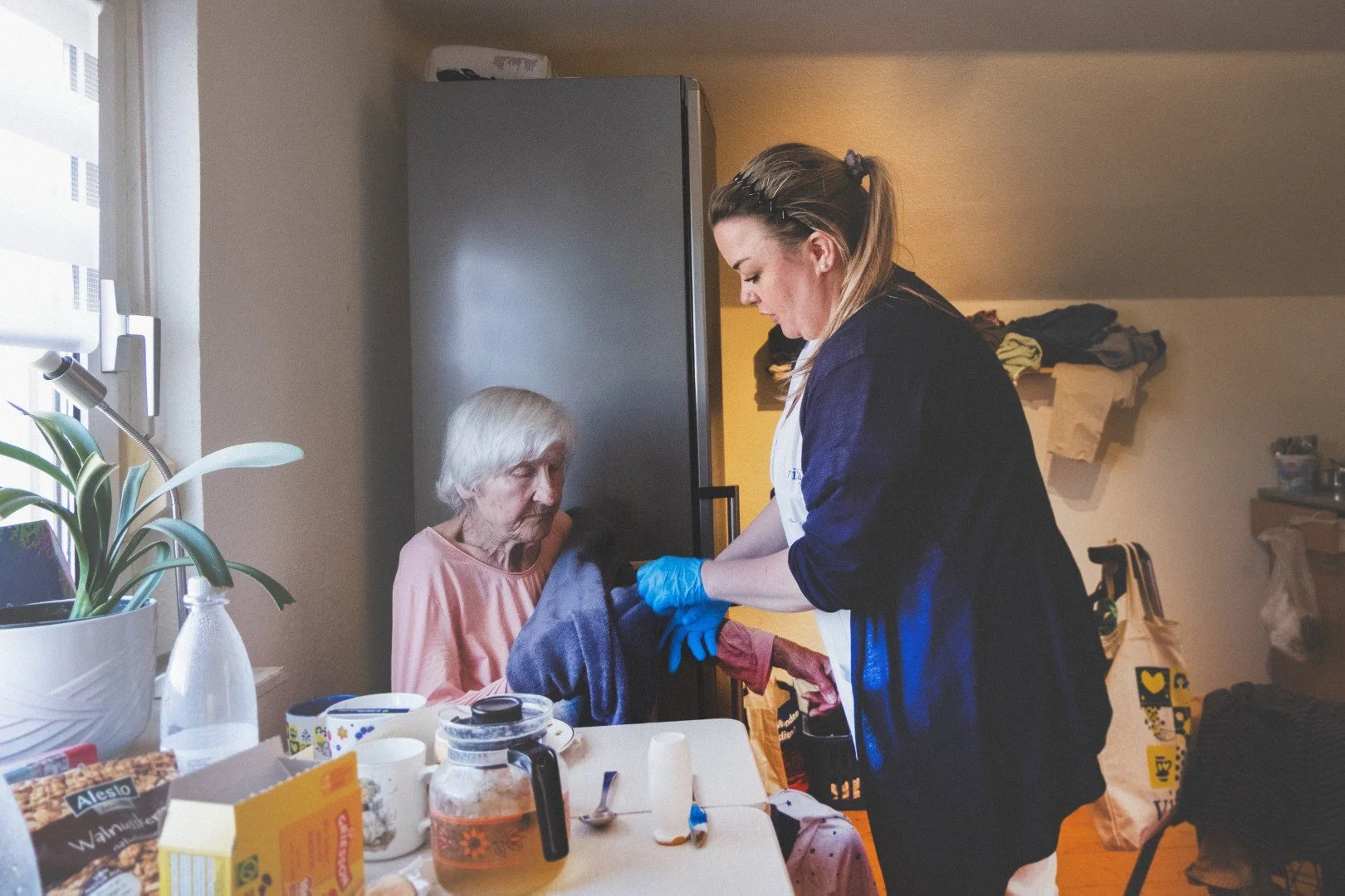 Pflegekraft versorgt eine ältere Patientin am Tisch in einem Wohnraum.