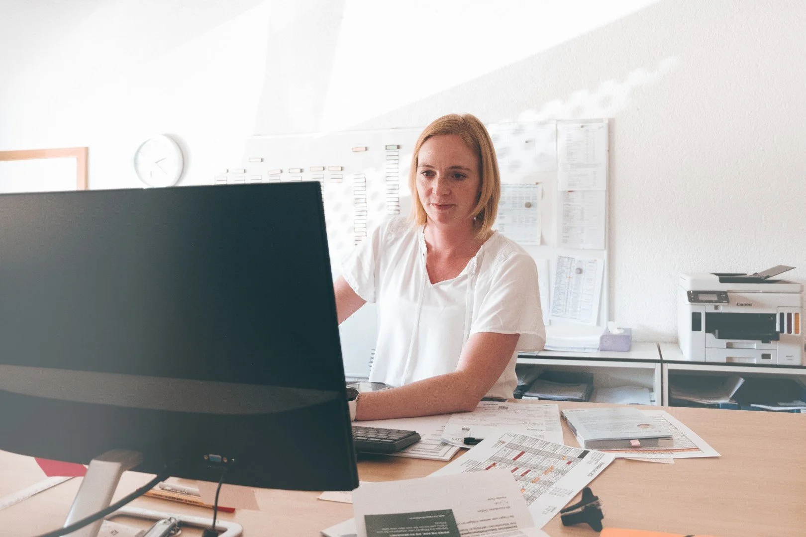 Frau arbeitet an einem Schreibtisch im Büro mit Computerbildschirm, Papieren und Drucker.