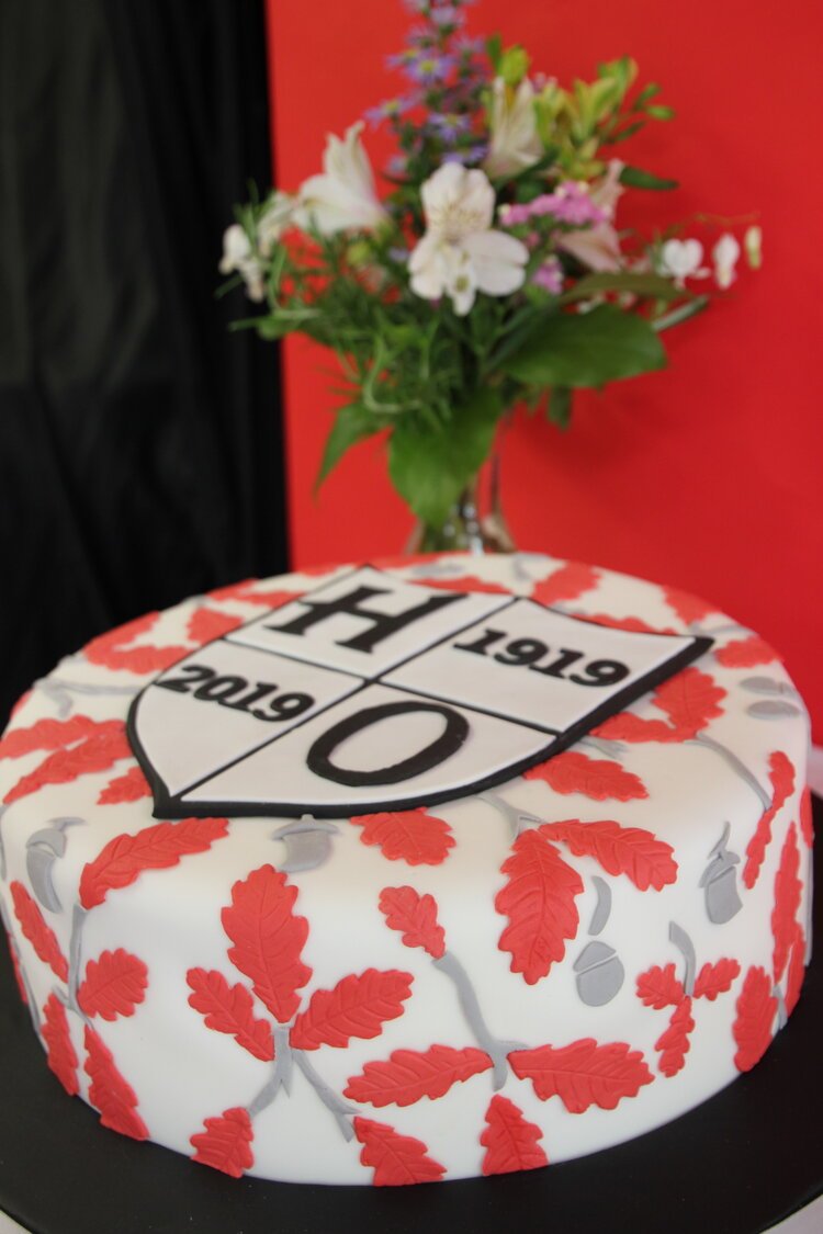 Decorative cake with red and grey leaf design, featuring a shield emblem marked "H 2019 1919," accompanied by a floral arrangement in the background.