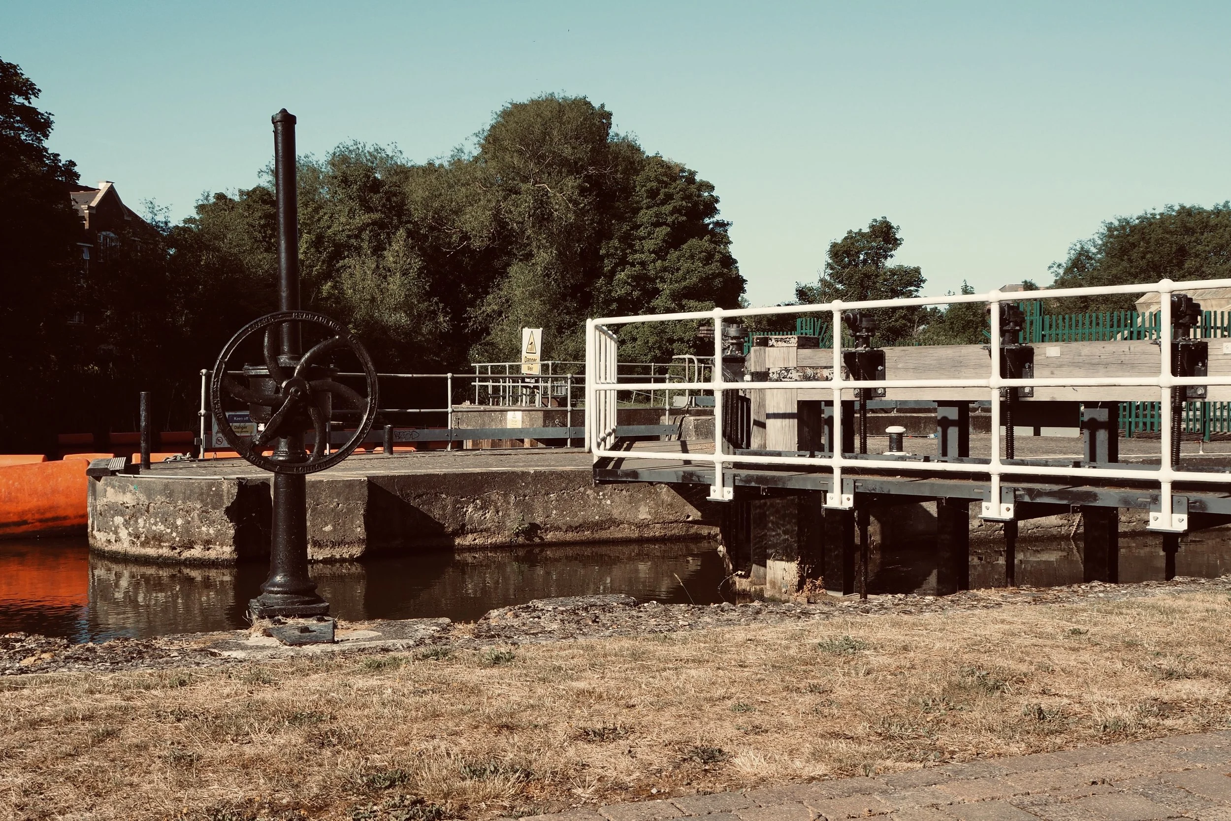 Canal lock with black metal wheel, white railings, and surrounding foliage on a sunny day.