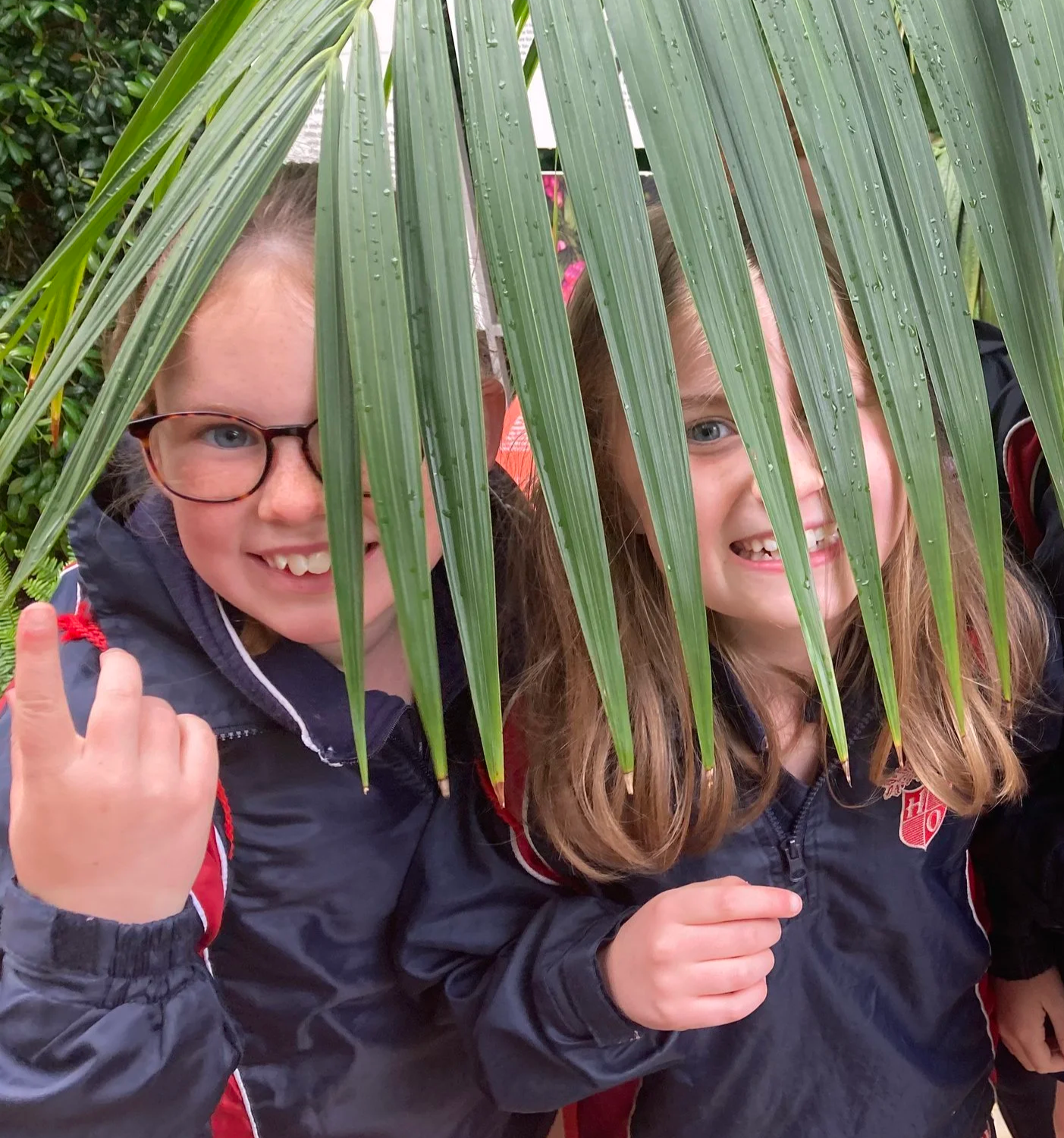 Three children smiling and posing behind a large green palm leaf, outdoors, wearing navy jackets with red accents.