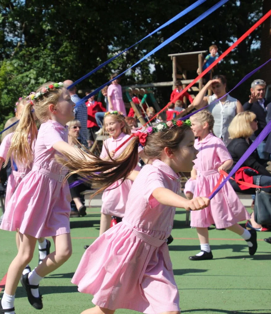 Children in pink dresses participating in a traditional Maypole dance outdoors during a sunny day, with spectators in the background.