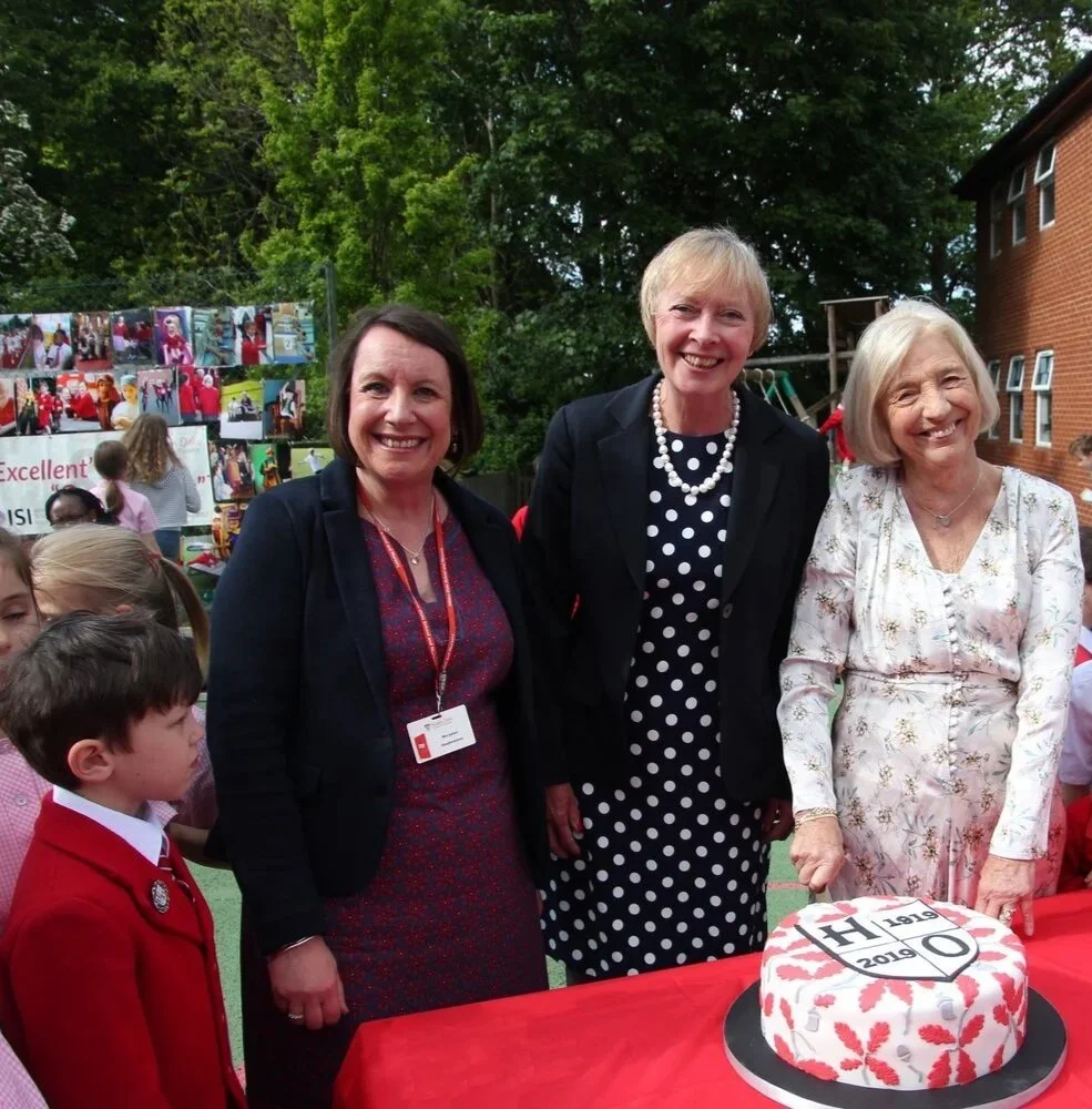 A group of people, including three women and several children, gathered around a table with a red cloth, featuring a decorative cake with the years 1879-2019 in an outdoor setting.