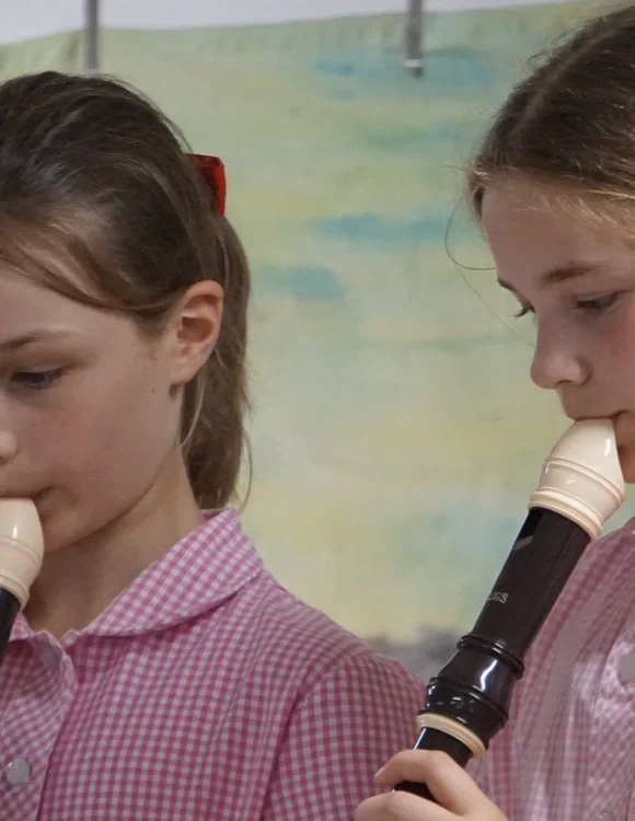 Two young girls in pink checkered shirts playing recorders together, focusing on their music.