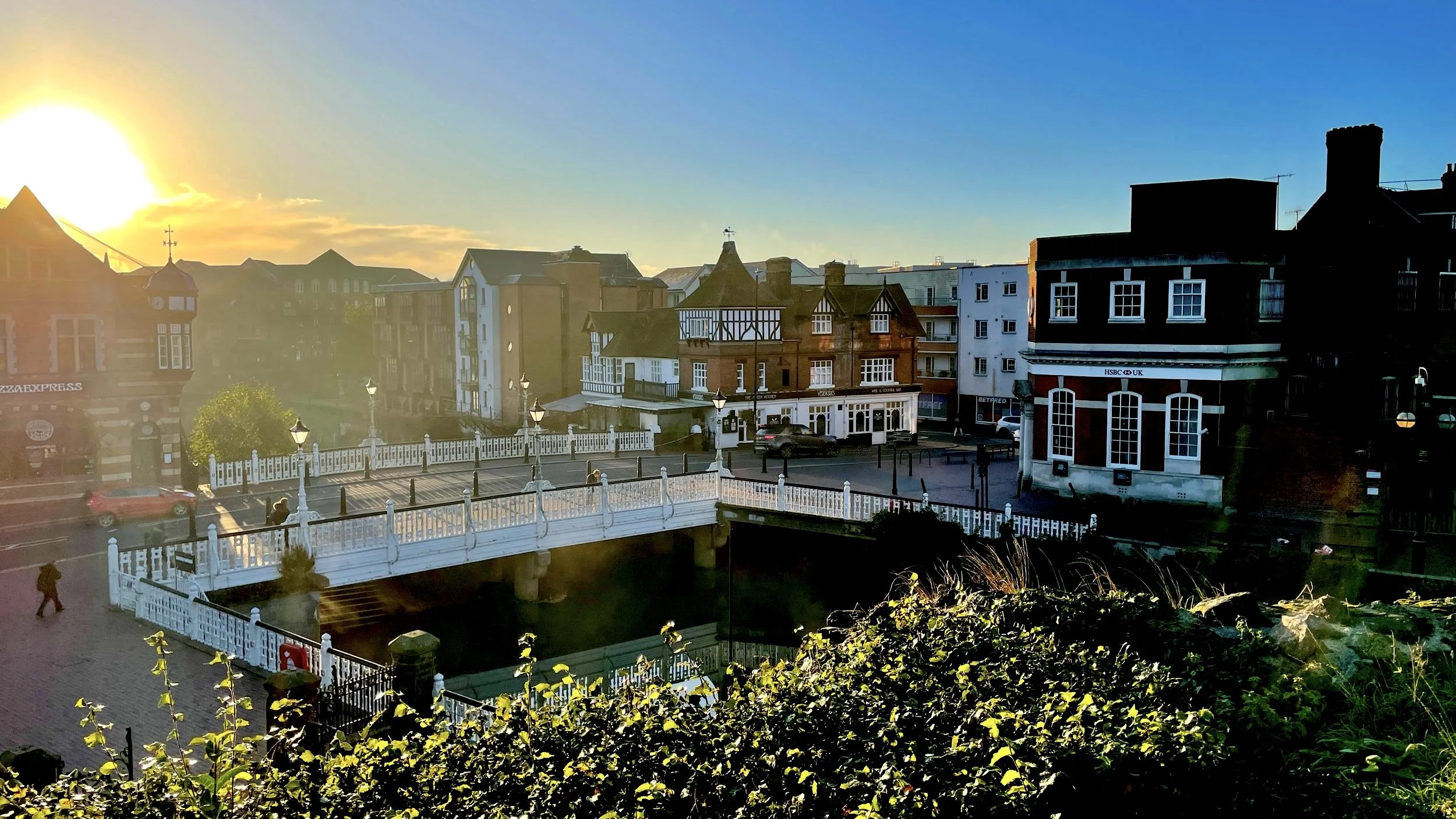 A scenic view of a historic town with a prominent bridge, traditional architecture, and a sunlit sky. The foreground features a bridge with white railings, while the background includes brick and half-timbered buildings. The sun is setting on the horizon, casting a warm glow over the scene.