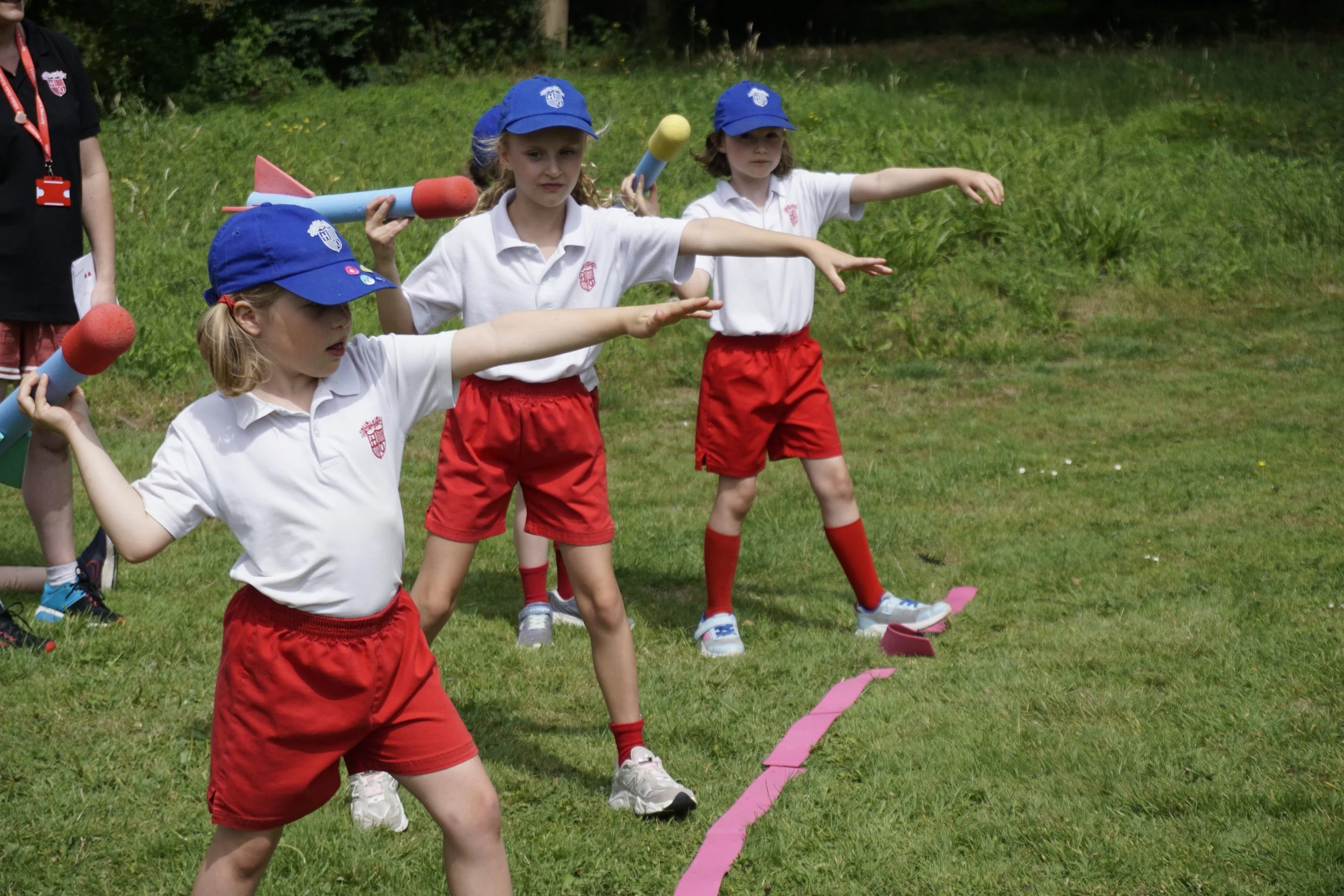 Young children dressed in white shirts, red shorts, and blue hats participating in an outdoor game, each holding foam rocket launchers, standing on grass with a pink line marked on the ground, with trees in the background.
