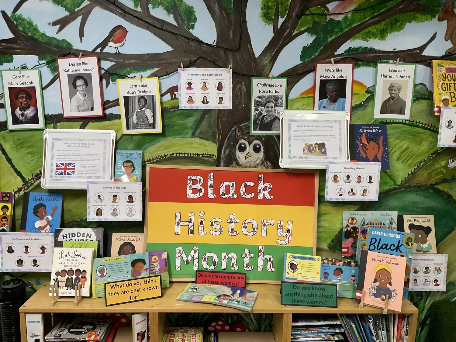 A colorful display celebrating Black History Month with books, photos, and information about influential Black women, set against a painted outdoor scene with trees and animals.