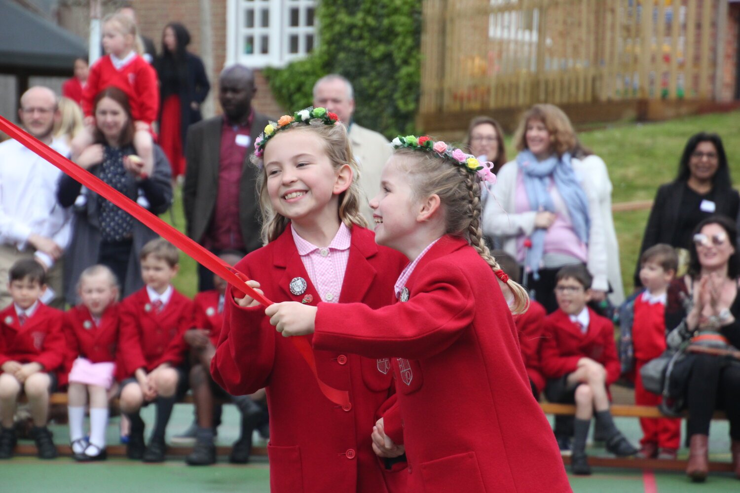 Two young girls in red uniforms with flower crowns smiling and holding a ribbon, surrounded by seated and standing people.