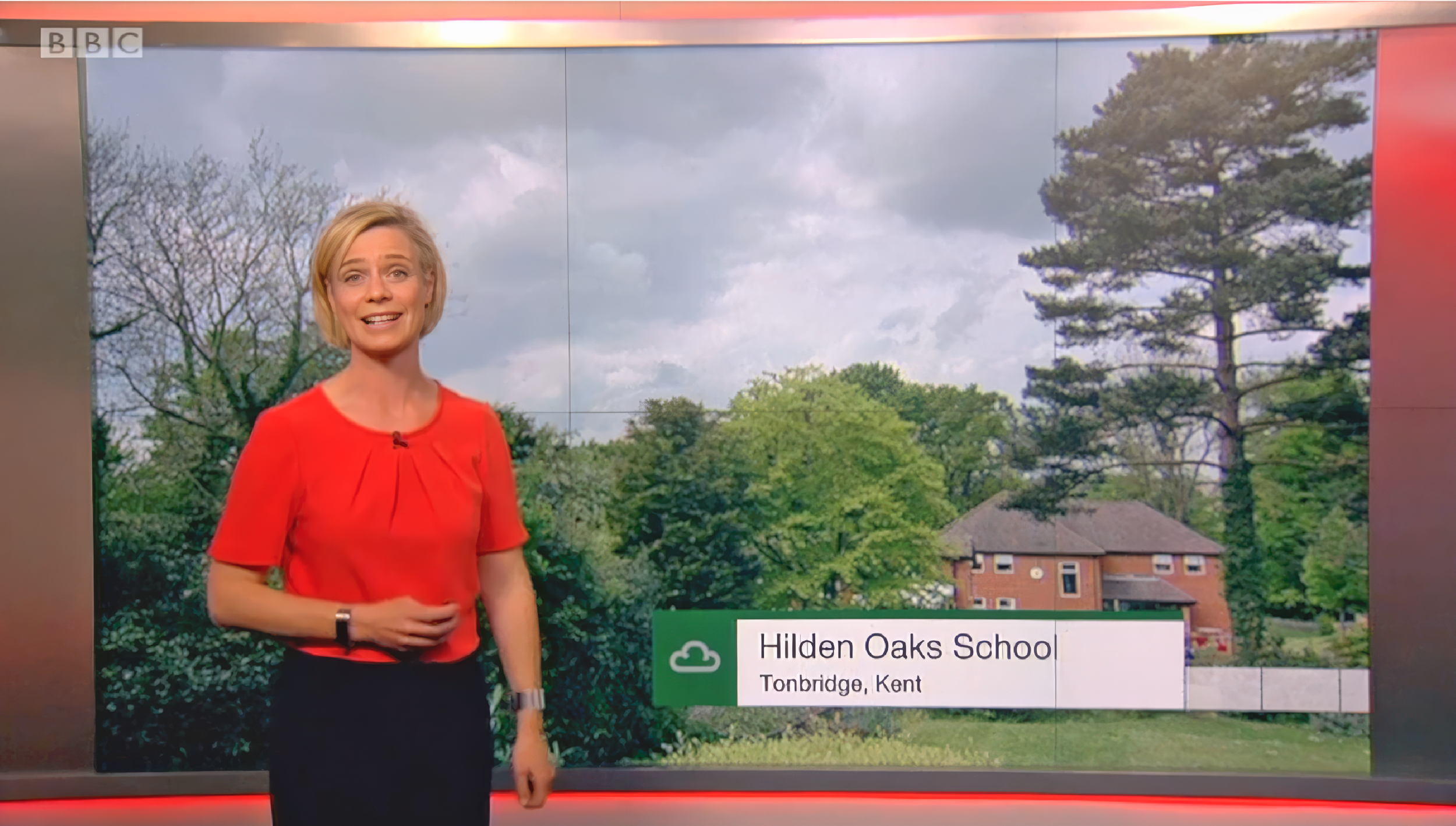 Weather presenter in a red dress stands in front of a screen showing a cloudy sky and trees with a location caption "Hilden Oaks School, Tonbridge, Kent."