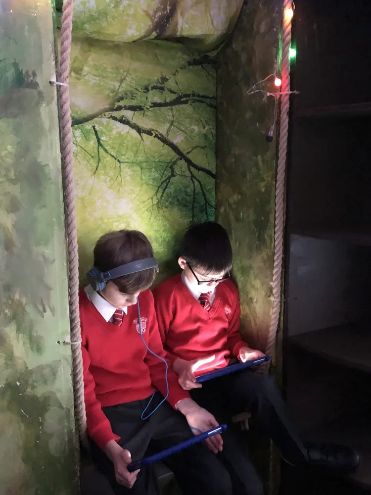 Two boys in red school uniforms sitting in a small, enclosed space decorated with a forest mural, using tablets and listening with headphones and glow sticks.