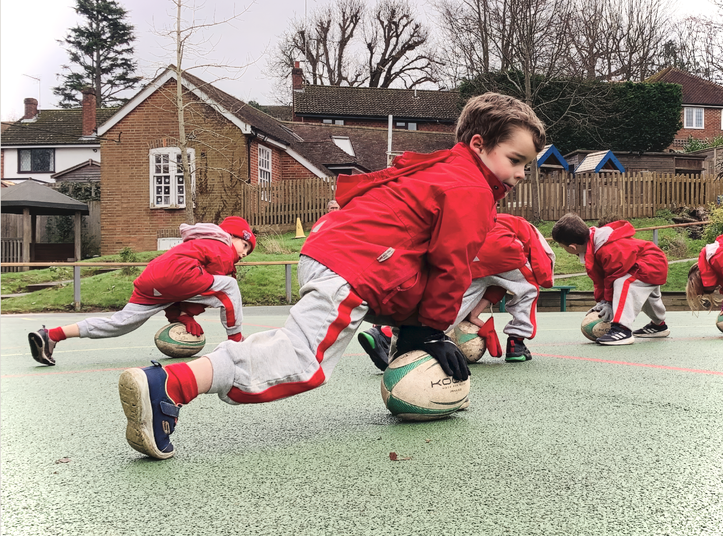 Young children in red jackets and gray sweatpants participating in a rugby drill on a playground, each crouching with one foot on a rugby ball, outdoors in a residential area with houses and trees in the background.