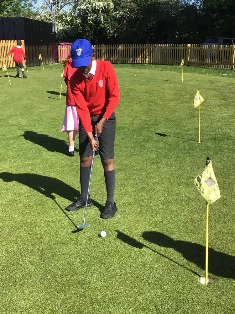 A child in a red school uniform and black shorts is practicing putting on a green putting green with small yellow flags. The child is wearing a blue cap and black shoes, and is focused on lining up a golf putt. In the background, another child in a red uniform is also on the green, and there is a wooden fence with trees behind it.