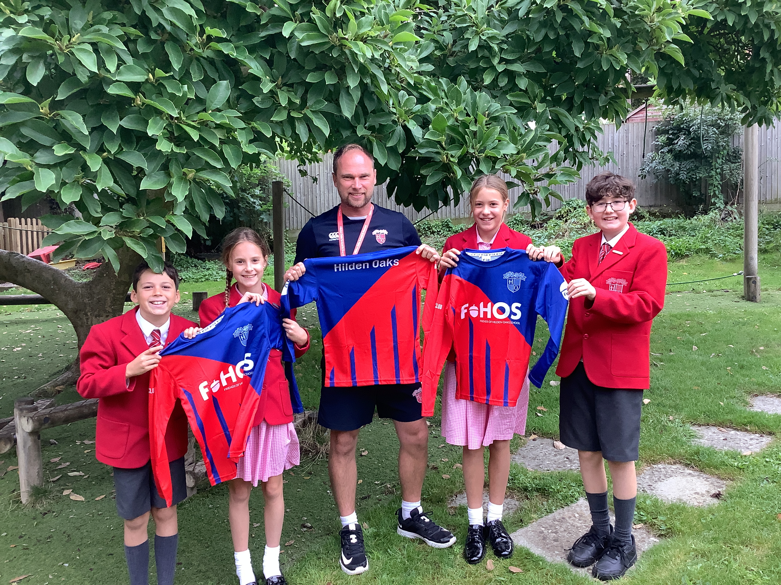 A man and four children in school uniforms standing outdoors under a tree, holding rugby jerseys with