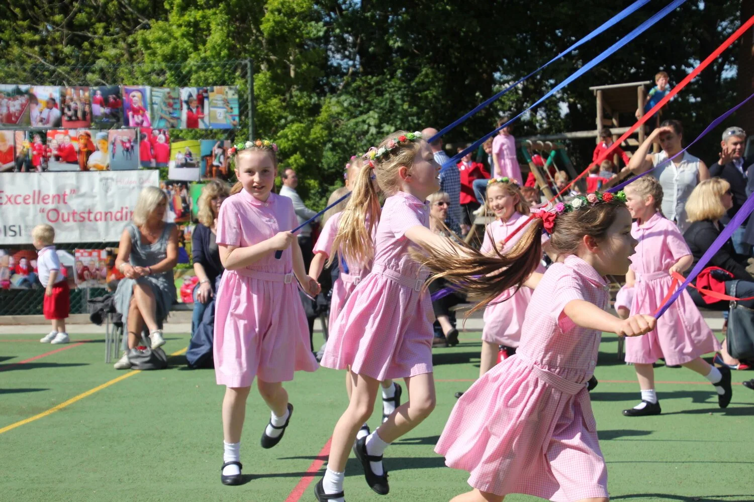 Children in pink dresses participating in a traditional Maypole dance outdoors during a sunny day, with spectators in the background.
