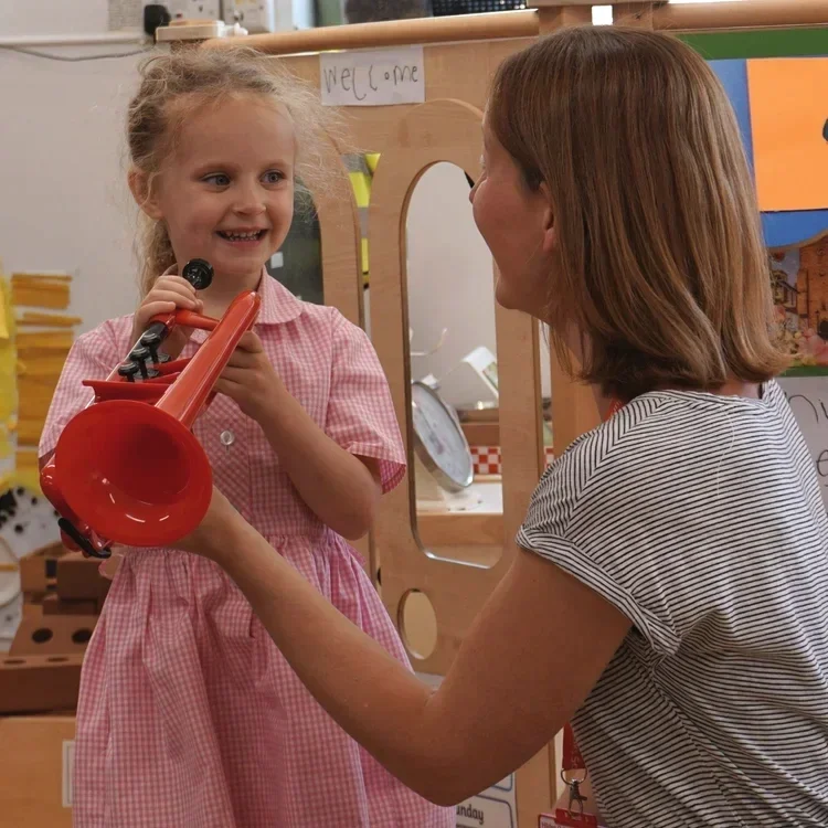 A young girl in a pink checkered dress smiling as she holds a red toy trumpet, while a woman with short brown hair in a striped shirt holds the trumpet towards the girl, in a classroom or playroom setting.