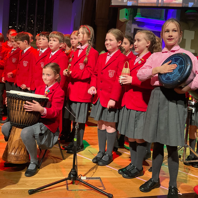A group of school children in red blazers, gray skirts or pants, standing on stage during a performance. One girl is sitting and playing a large drum, while another girl holding a round drum or tambourine is on the right side of the image. They appear to be singing or preparing to sing.