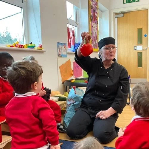 A woman in black scrubs and a black head covering holds a red object, possibly a toy, in front of a group of young children wearing red sweaters. The woman appears to be explaining or demonstrating something to the children in a classroom setting.