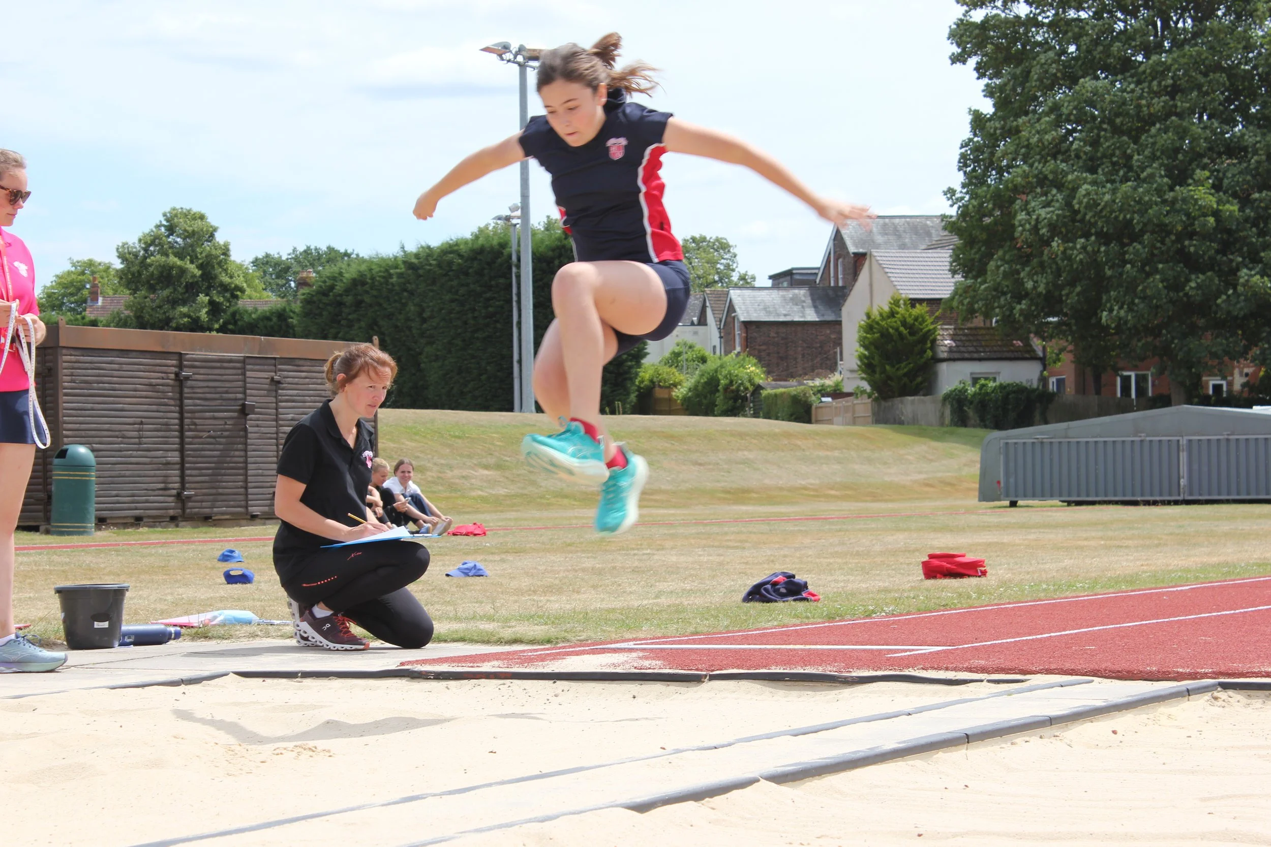 Female athlete in a black and red uniform performing a long jump at an outdoor track and field event with officials and spectators nearby.