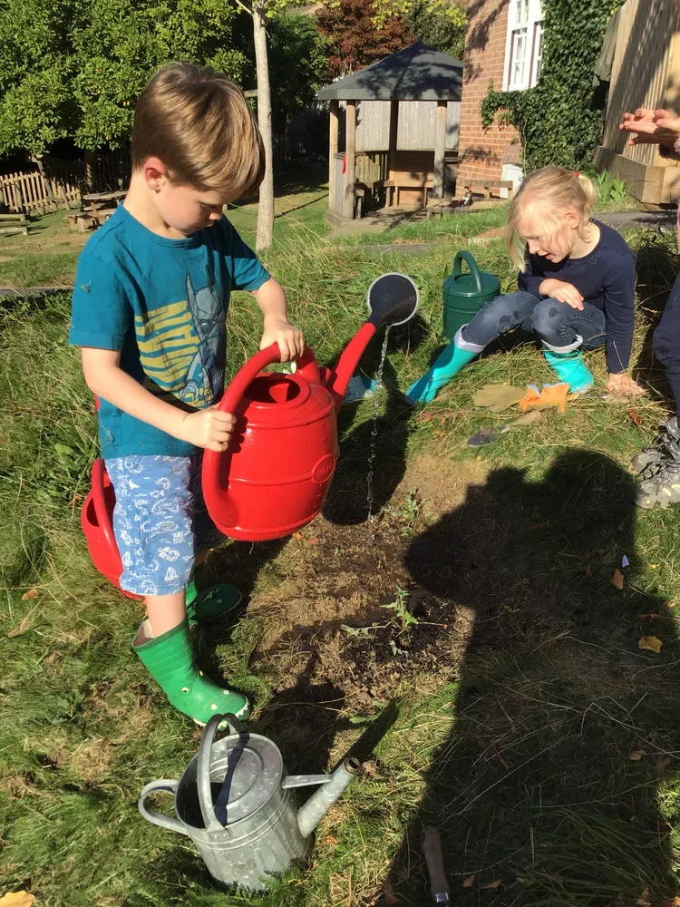 Two children watering plants in a garden with watering cans, one boy in green rain boots and one girl in turquoise rain boots, surrounded by gardening tools and greenery.