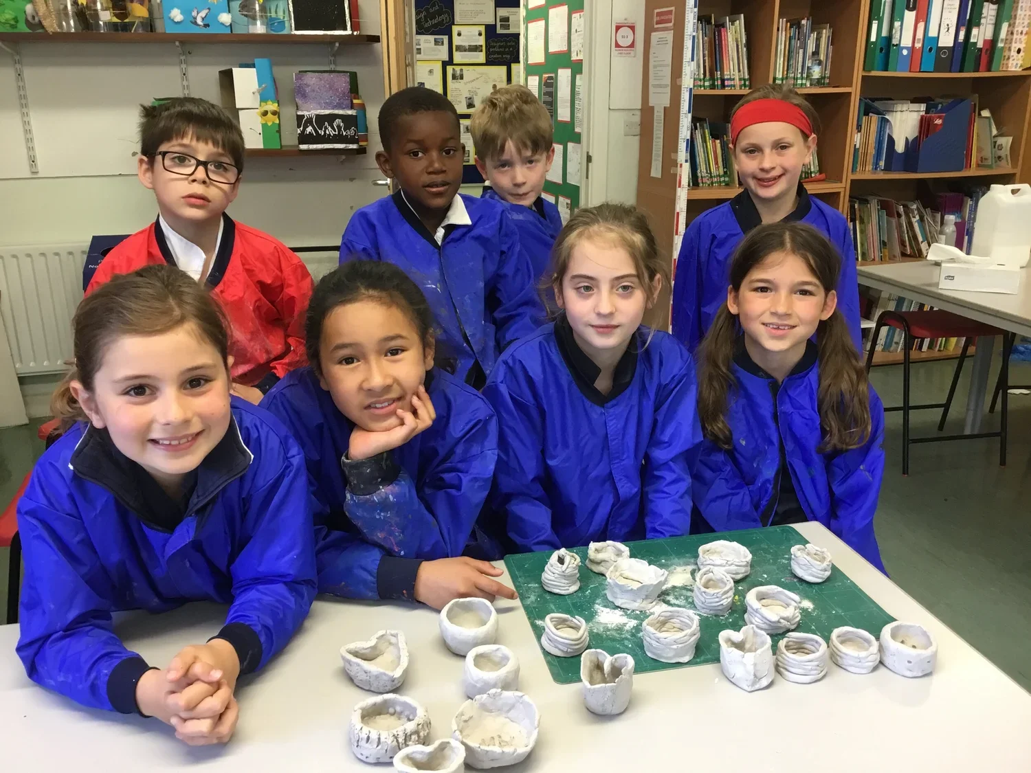 Group of children in blue smocks displaying pottery projects on a table in a classroom.