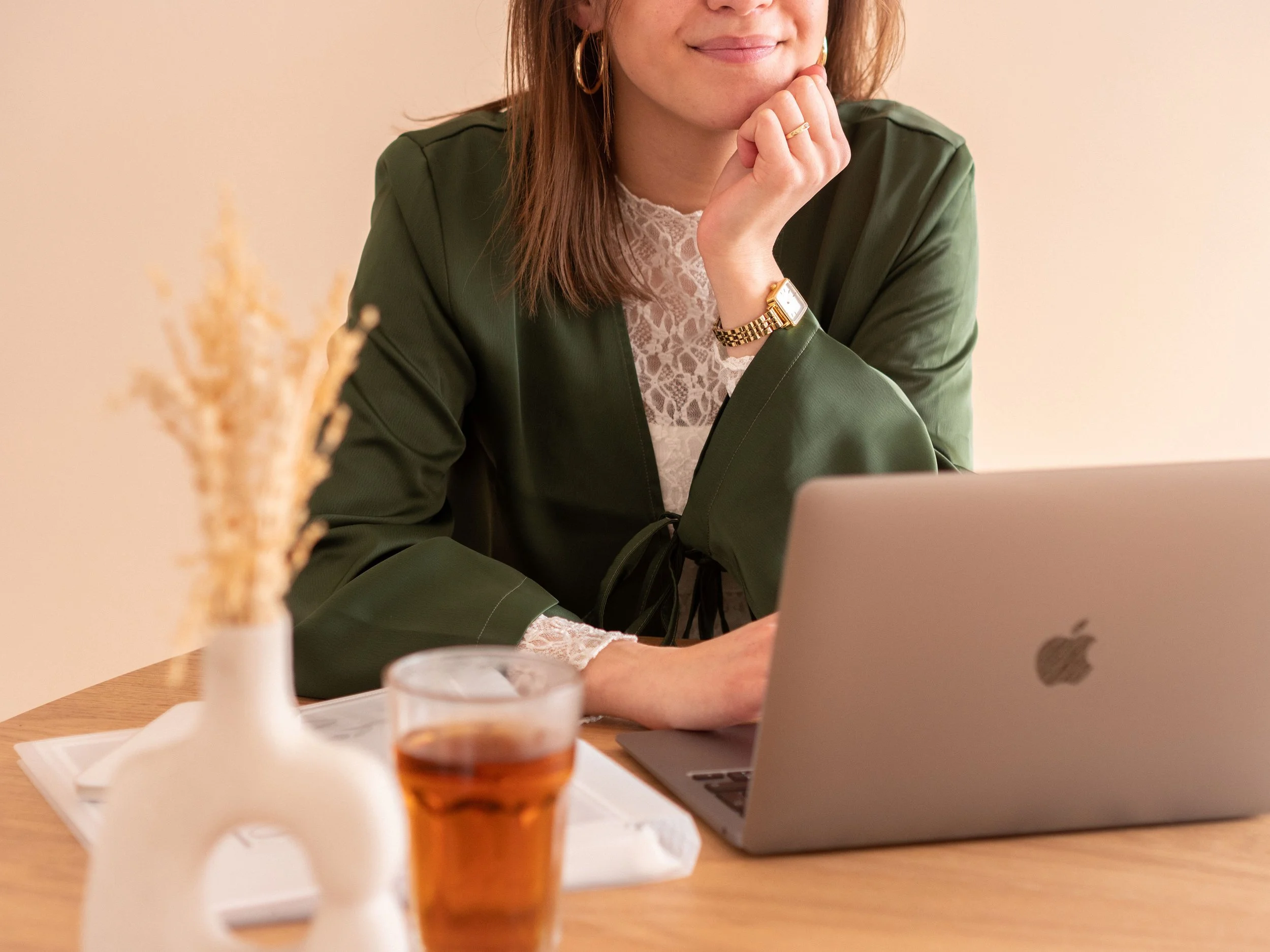 A woman with long hair, wearing a green jacket and a lace top, is sitting at a table with a laptop, a glass of tea, and a vase with dried flowers.