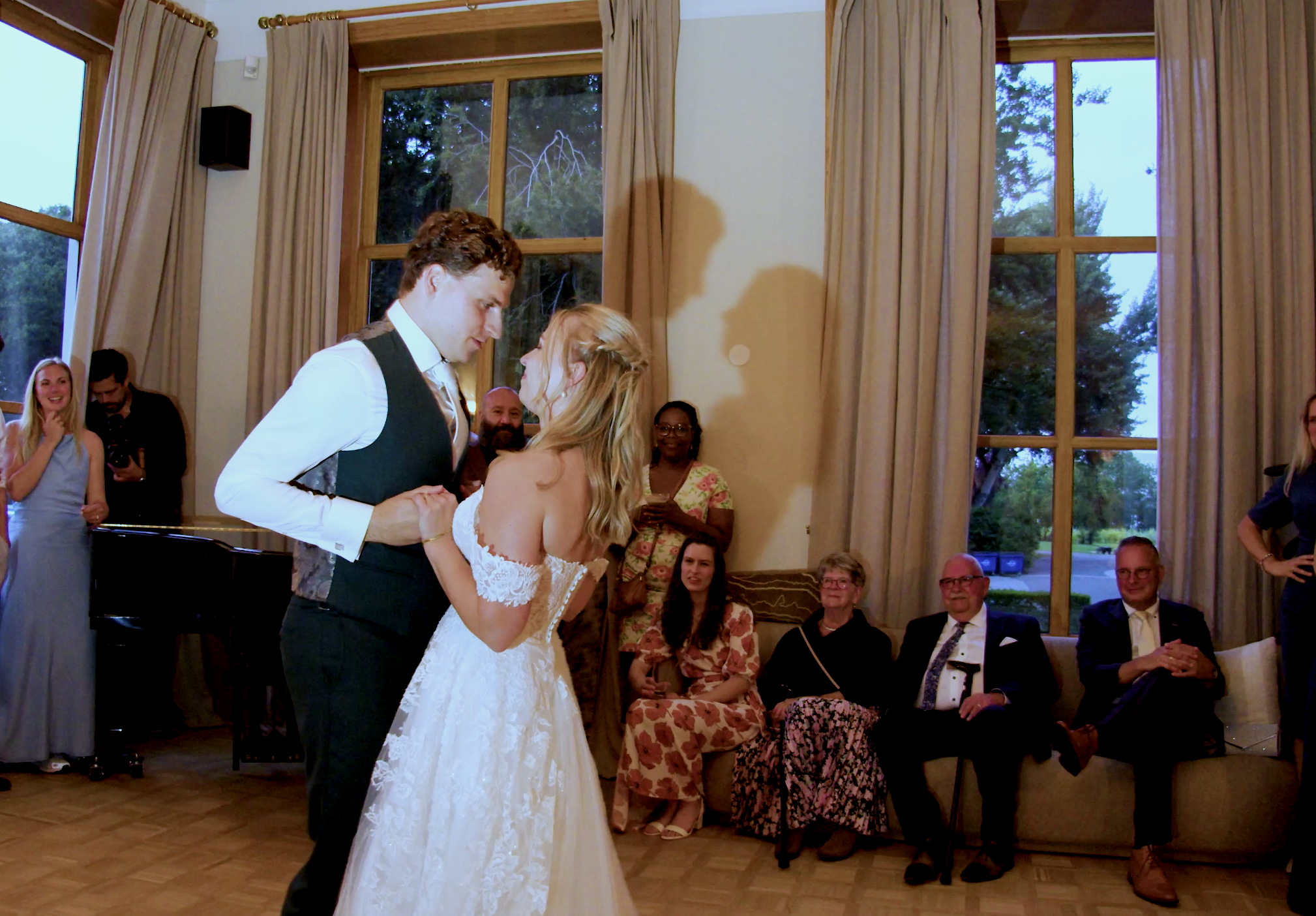 A bride and groom dance closely at their wedding reception with guests watching indoors near large windows.