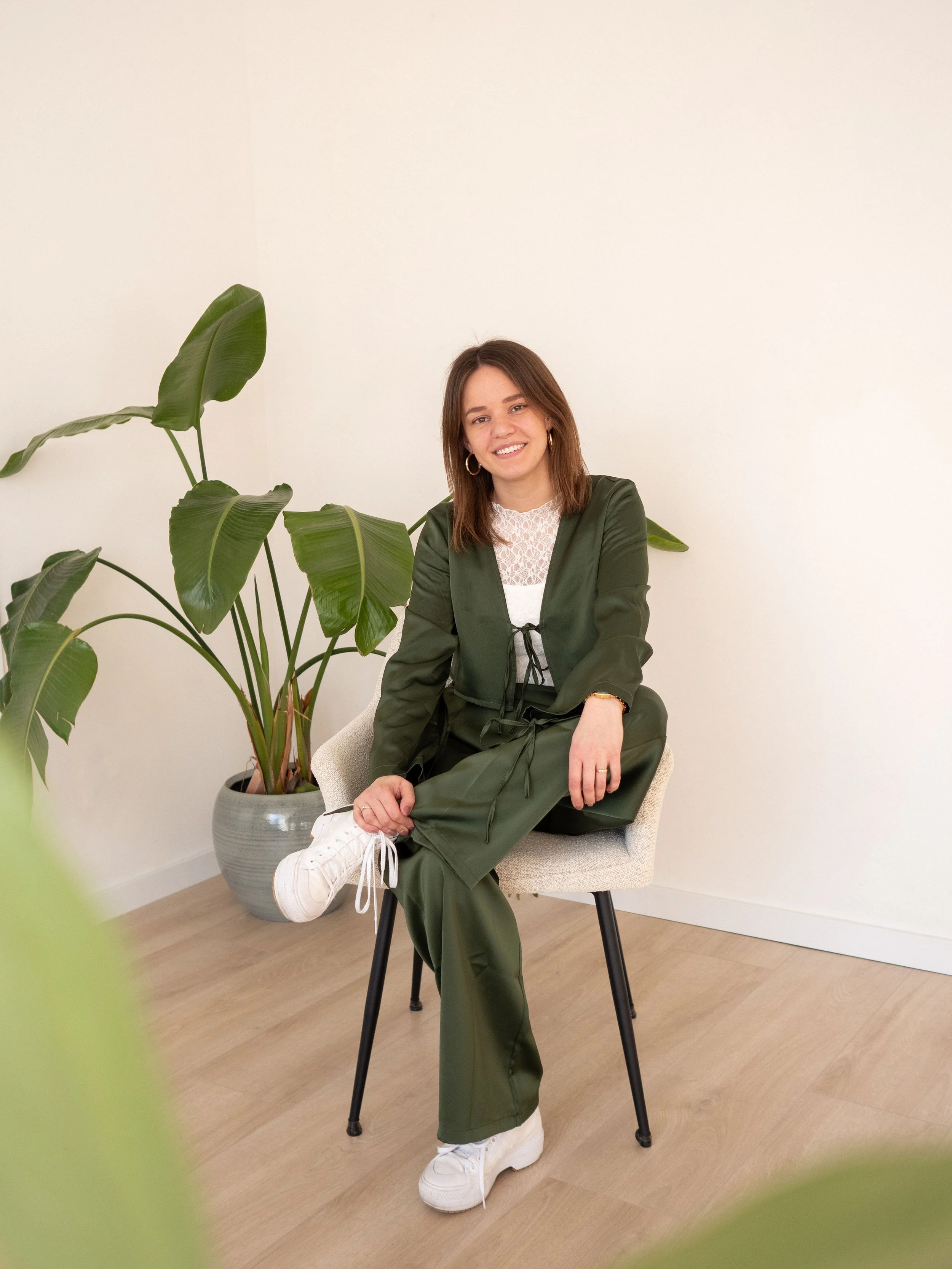 A woman with shoulder-length brown hair and hoop earrings, wearing a green satin outfit and white sneakers, sitting on a white chair with black legs in a minimalistic room with white walls and wooden flooring, next to a large potted plant.