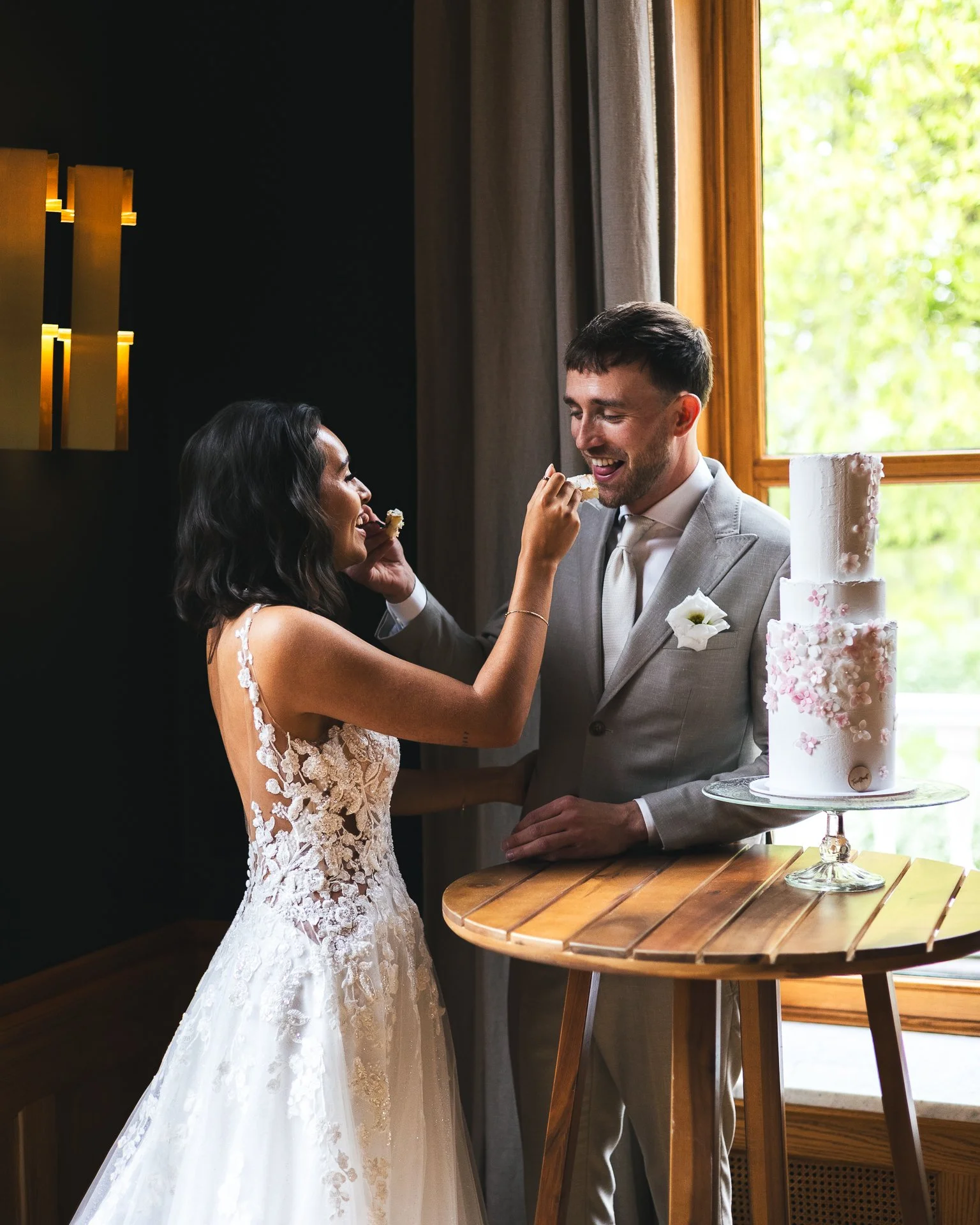 Bride feeding groom a slice of cake during wedding celebration, with a decorated wedding cake on a wooden table near a window.