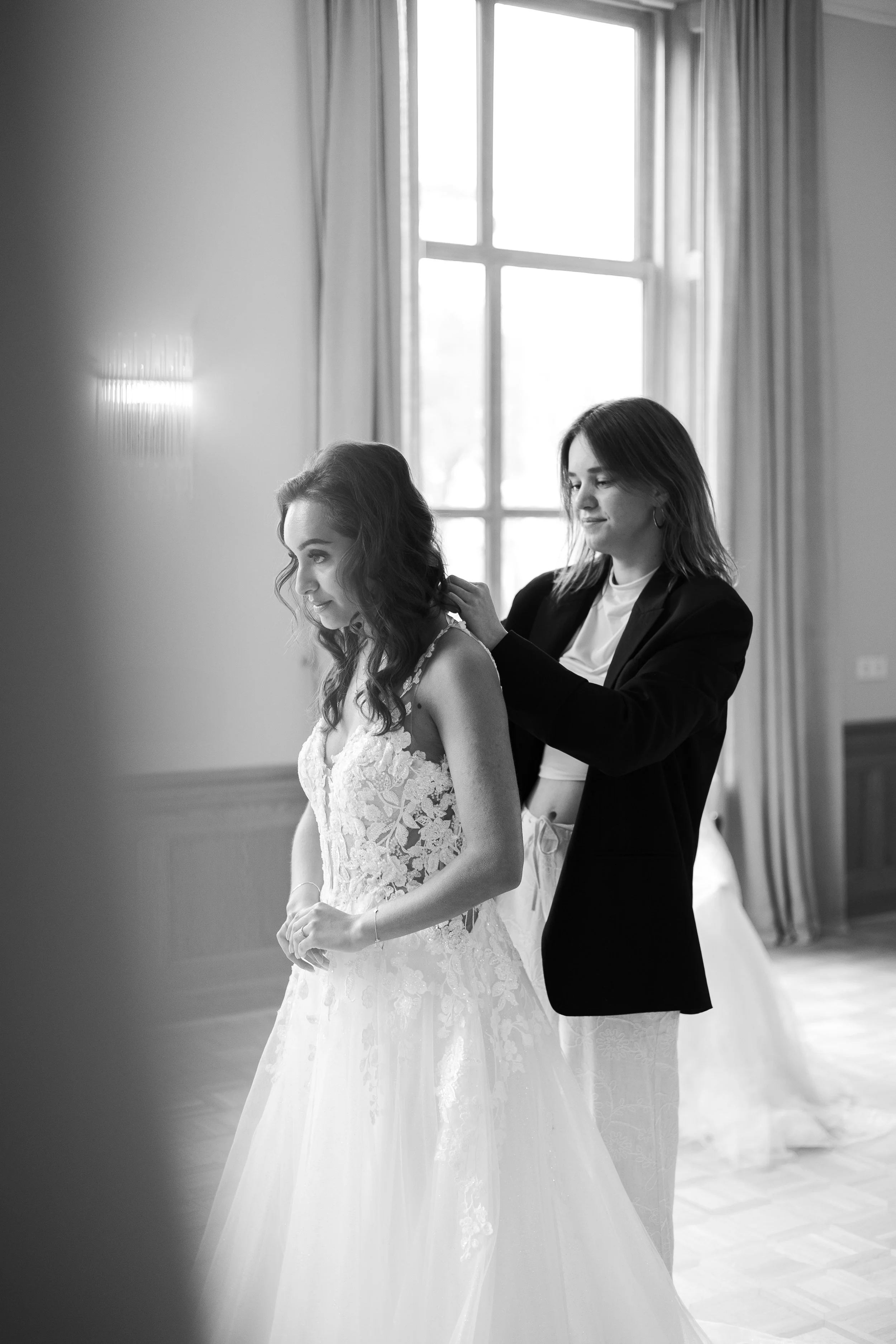 A woman in a lace wedding dress standing while another woman helps her with her hair inside a bright room with large windows.