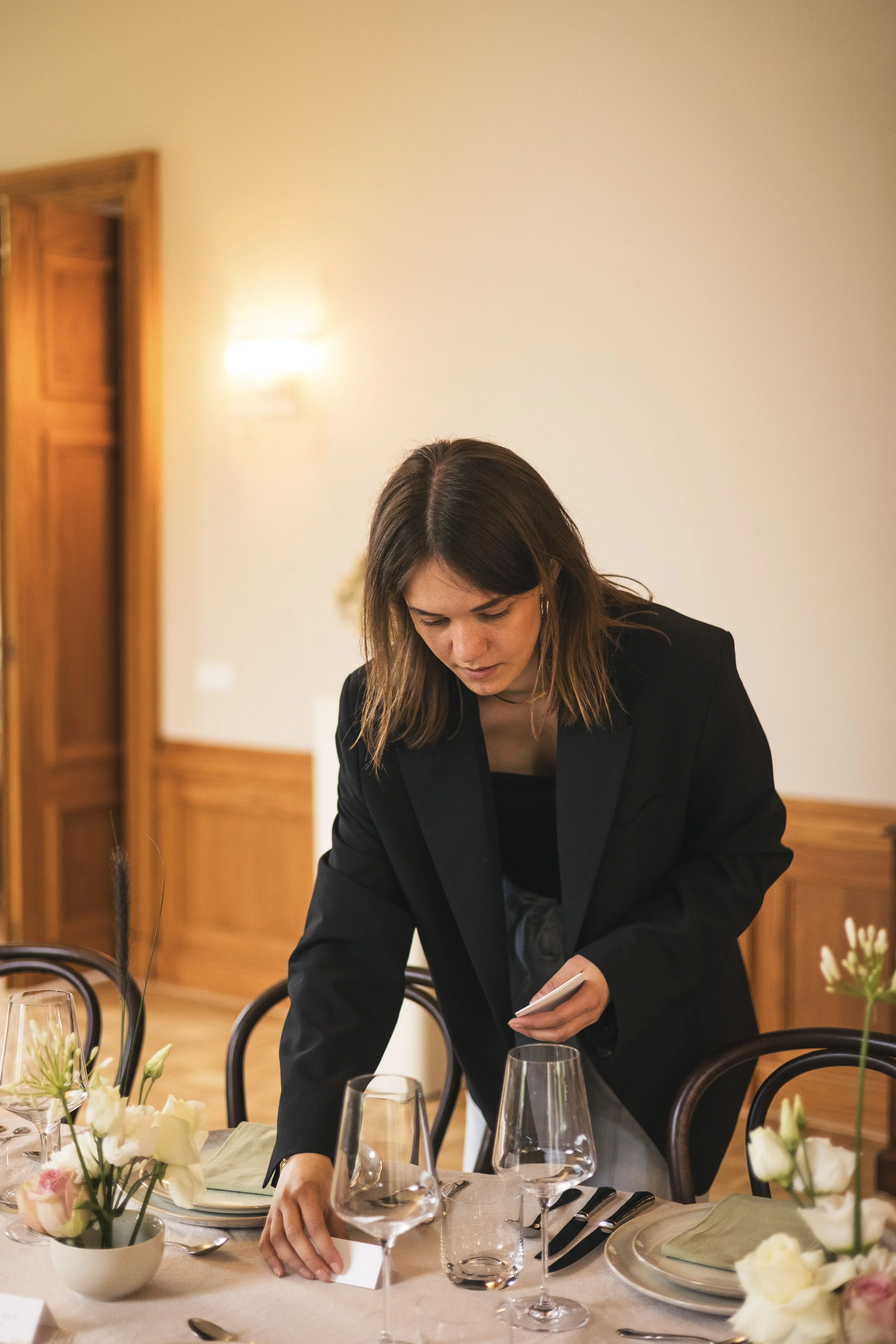 A woman in a black blazer arranging a dinner table with flowers, wine glasses, and plates.
