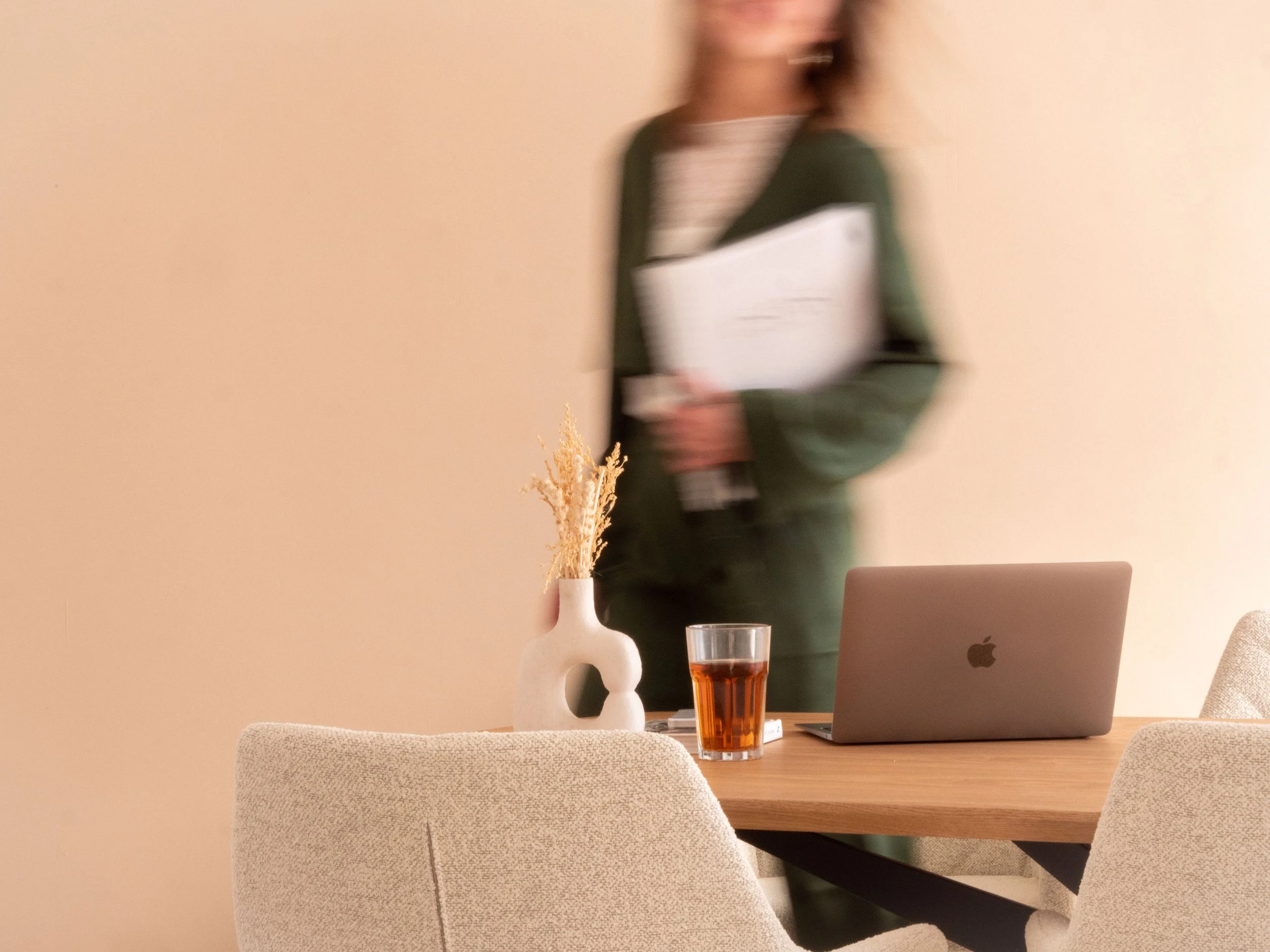 A blurred woman standing in a room holding a file folder, with a wooden table in the foreground containing a white vase with dried plants, a glass of iced tea, a closed laptop, and a small notebook.