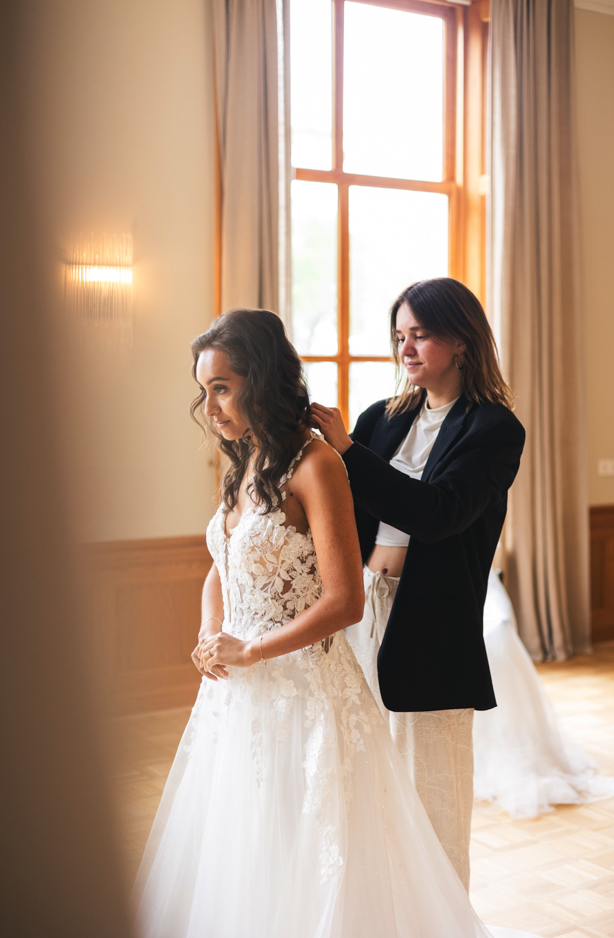 A woman in a lace wedding dress standing while another woman helps her with her hair inside a bright room with large windows.