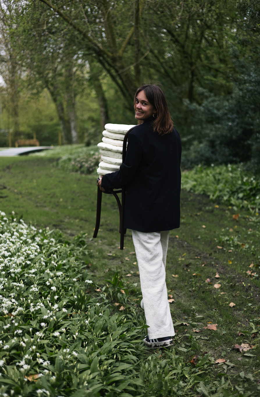Woman adjusting a wedding dress in a well-lit room with large window and ornate mirror.