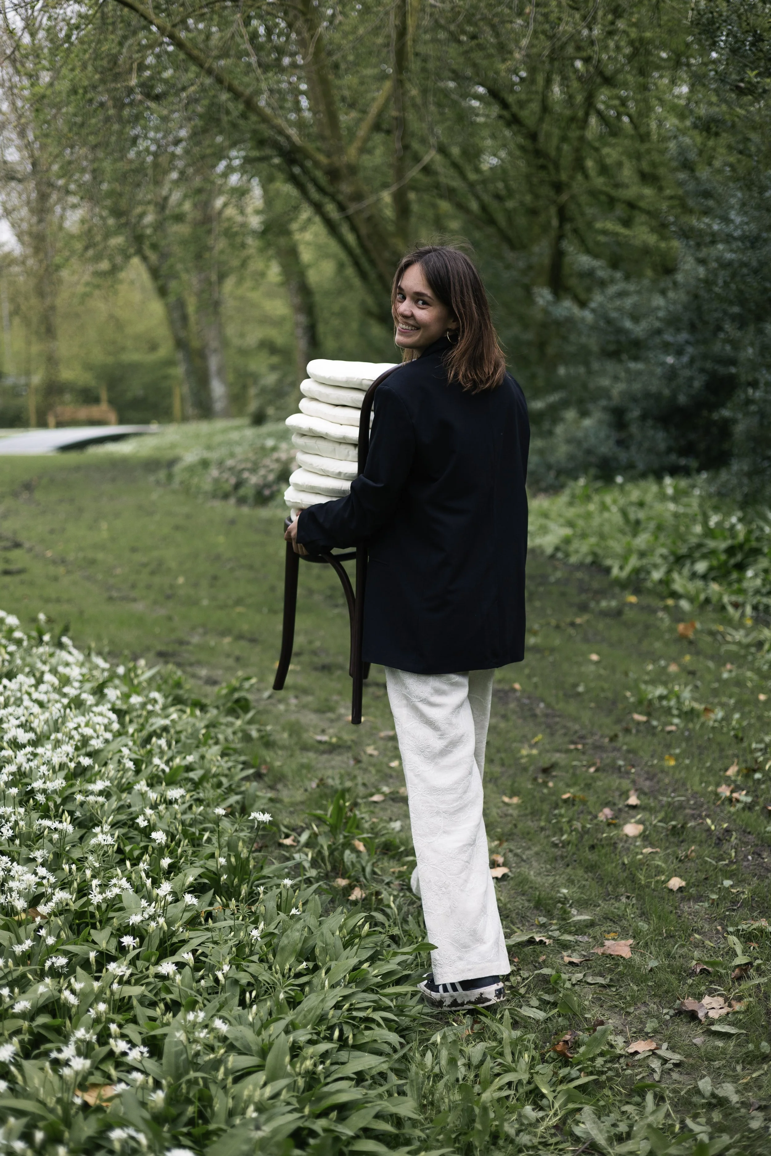 A young woman with shoulder-length brown hair smiles while walking outdoors on a path in a park with lush greenery and trees, carrying a stack of white towels or linens on a small stool.