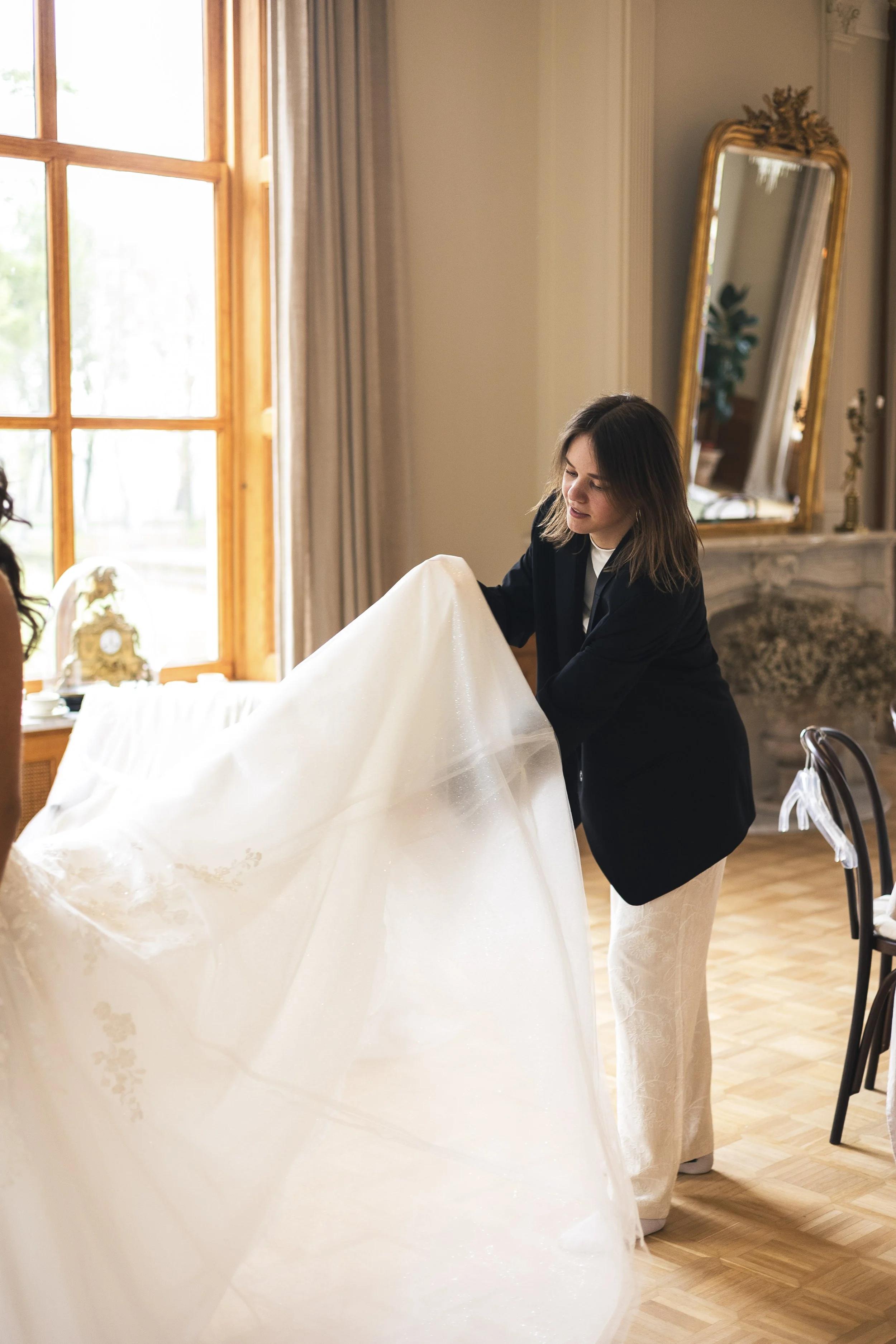 Woman adjusting a wedding dress in a well-lit room with large window and ornate mirror.