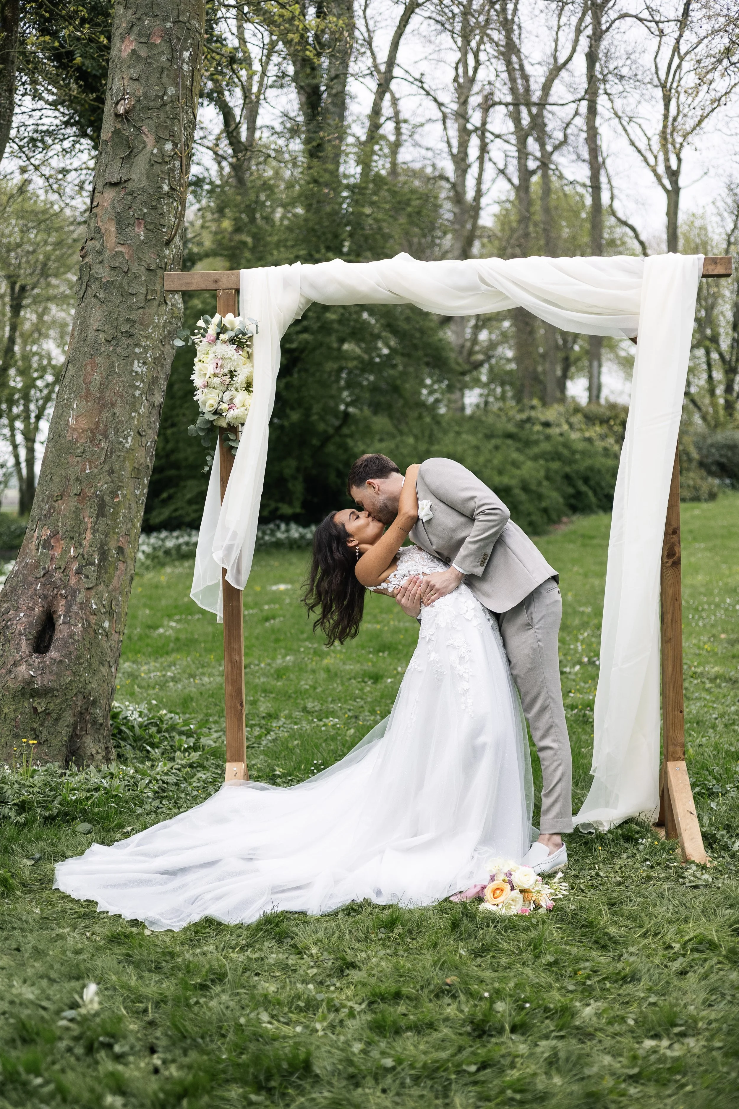 A newlywed couple sharing a kiss under an outdoor wedding arch during a spring or summer day, with lush green trees in the background.