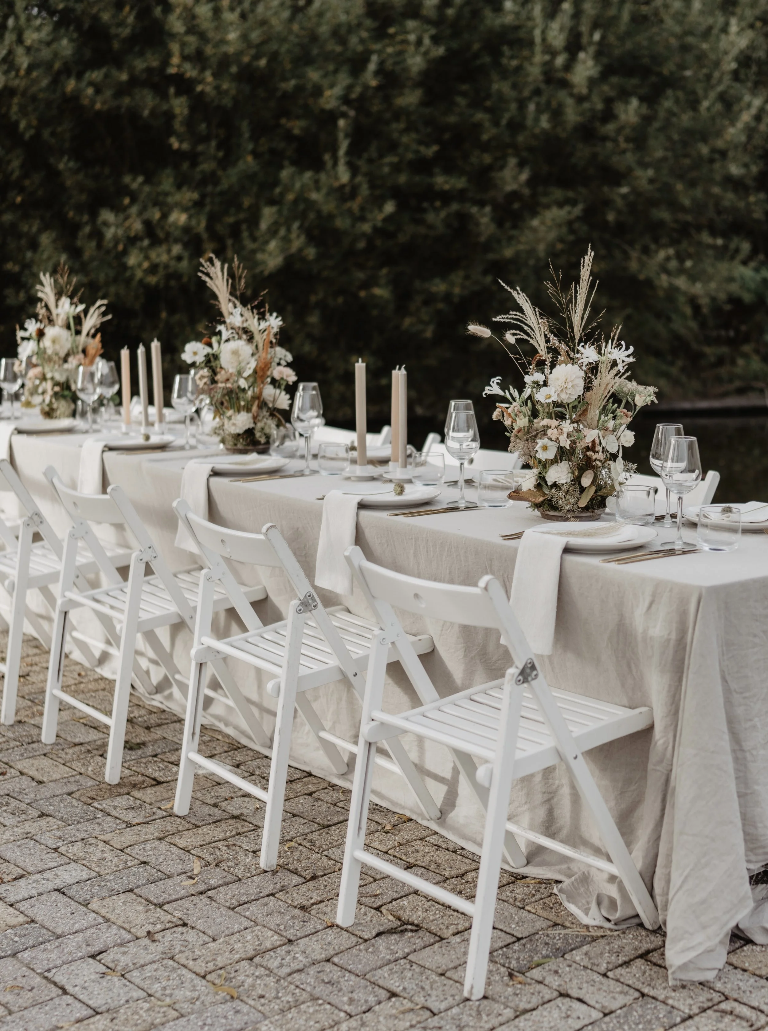Een lang diner tafel met een witte tafelkleed, bloemen en kaarsen buiten op een stenen vloer.