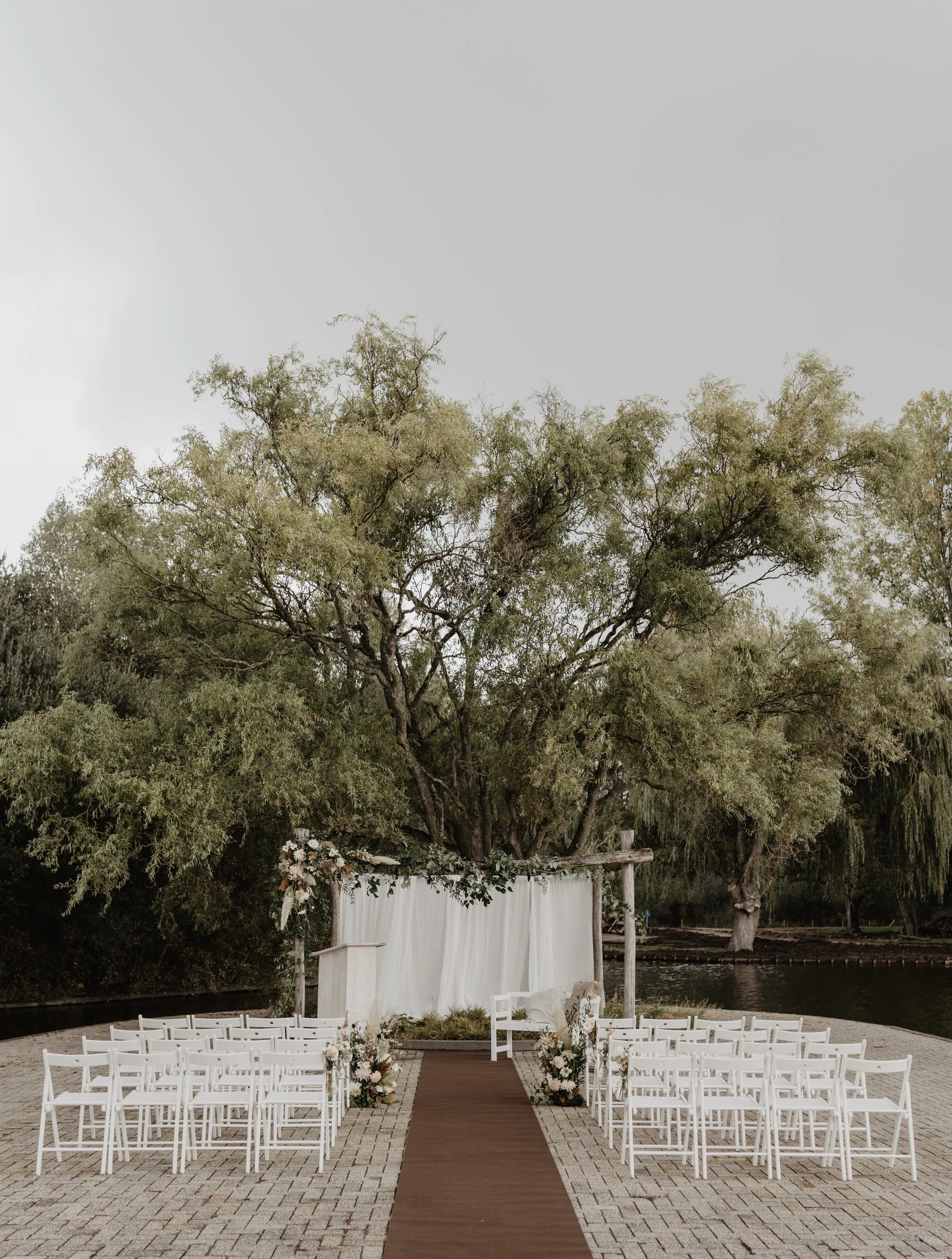 Een buitenceremonie met witte stoelen, een rode looplijn en een witte backdrops onder een grote boom naast een waterlichaam.