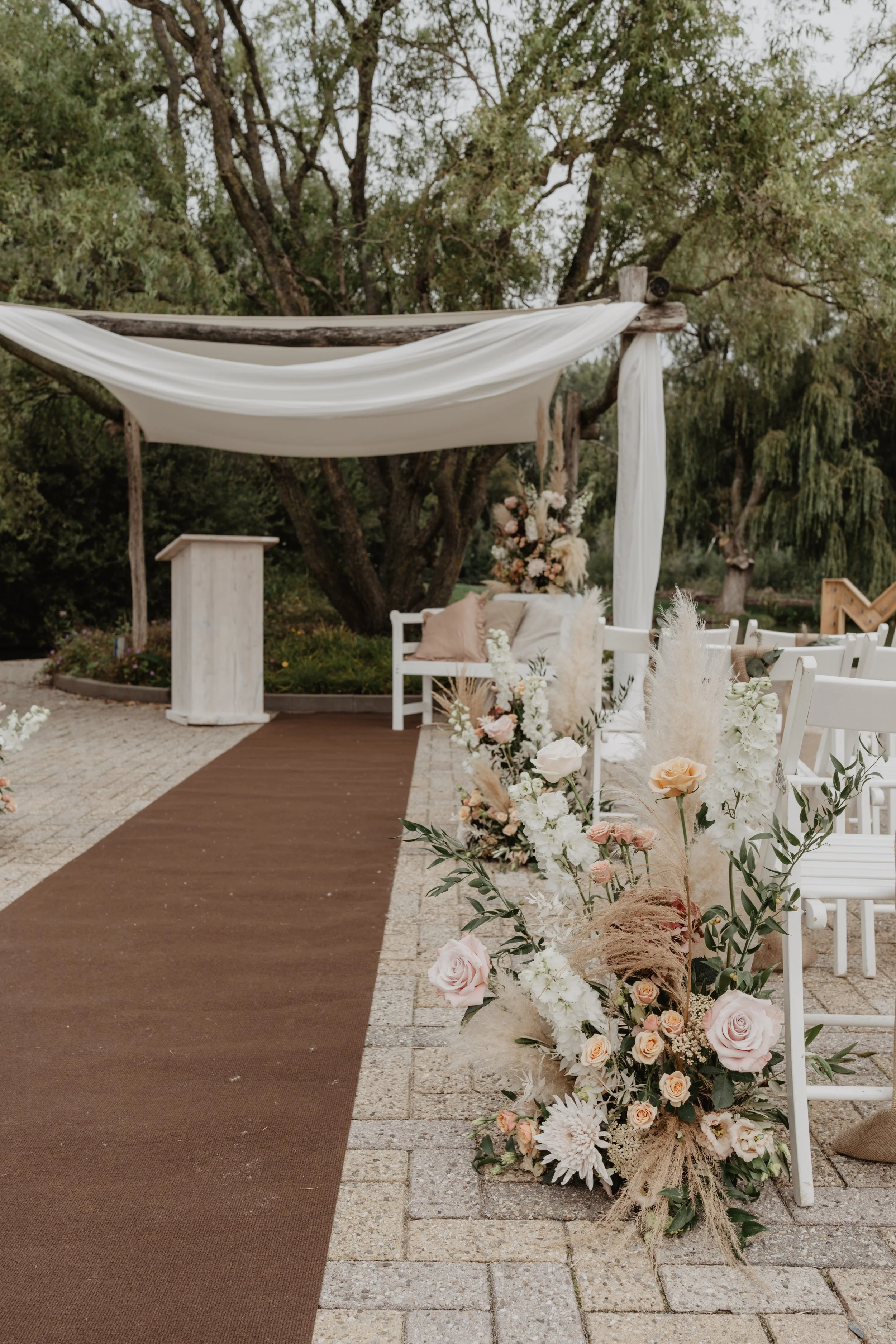 Decoratie voor een buitenceremonie met een bloemenkrans en een bank onder een houten structuur met witte stoffen draperie, in een tuinomgeving.