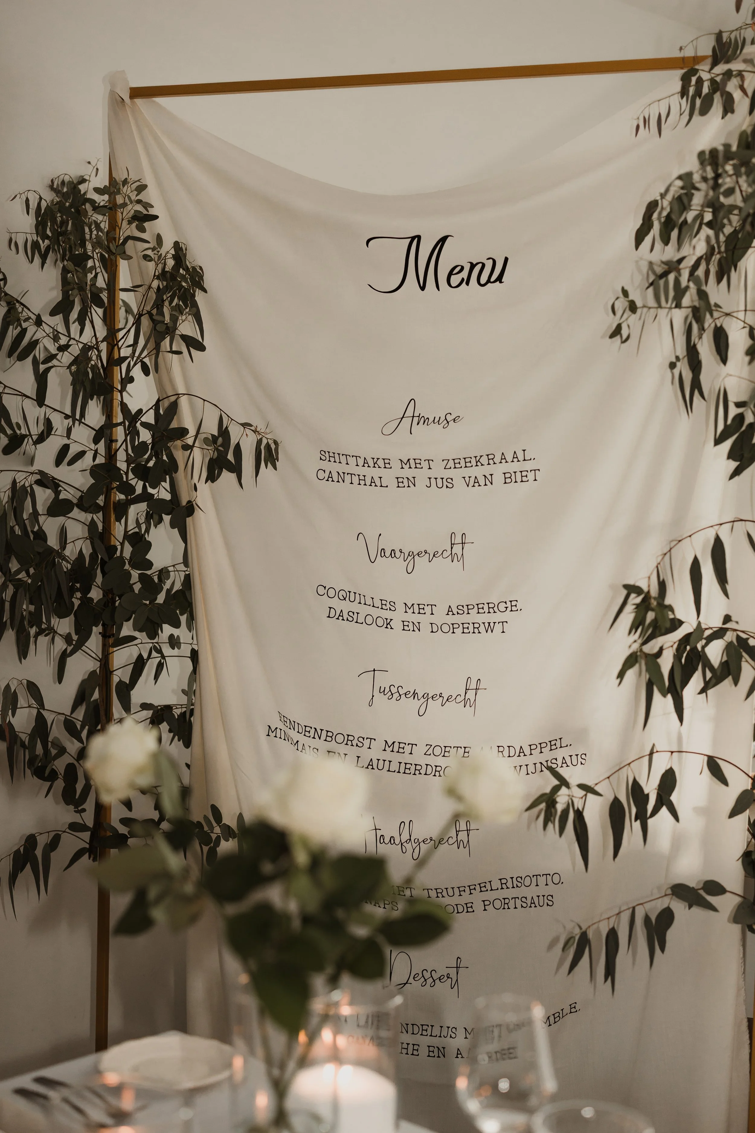 Menukaart met verschillende gerechten en categorieën, versierd met groene bladeren en witte bloemen op een gedekte tafel.