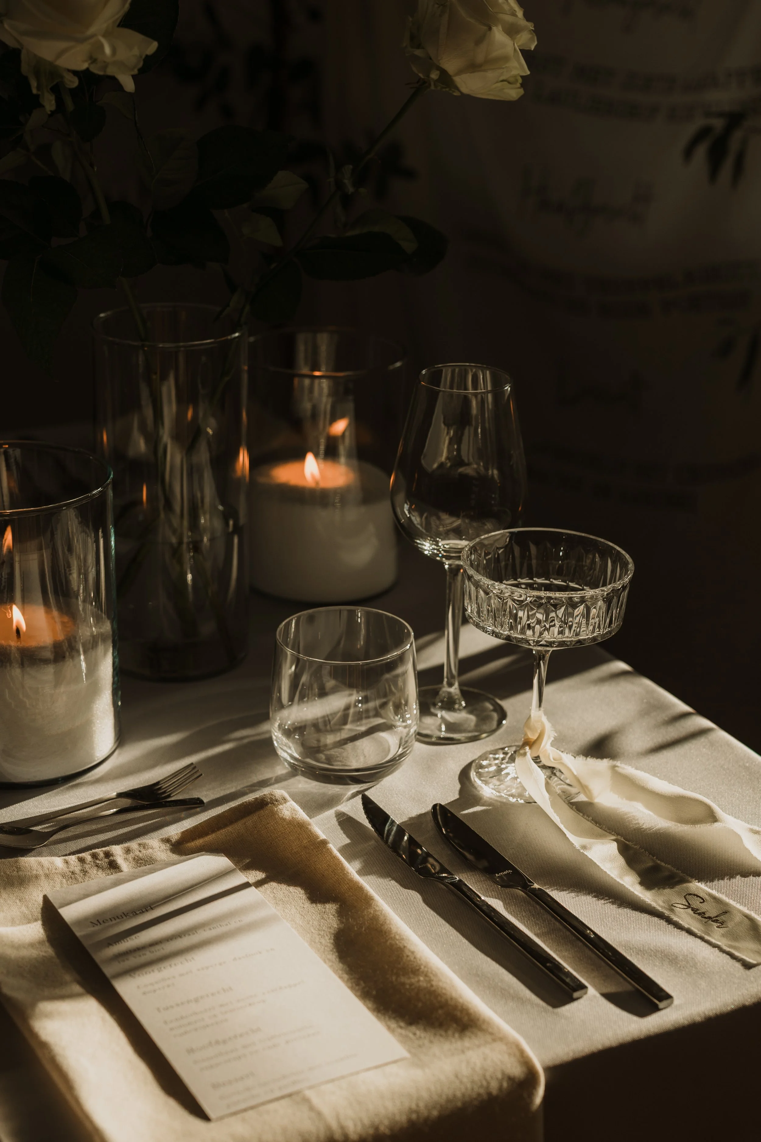 Elegante gedekte tafel met glazen, mes en vork, een menu, een kaars en witte rozen in een vaas, met sfeervol schijnsel en zachte schaduwen.