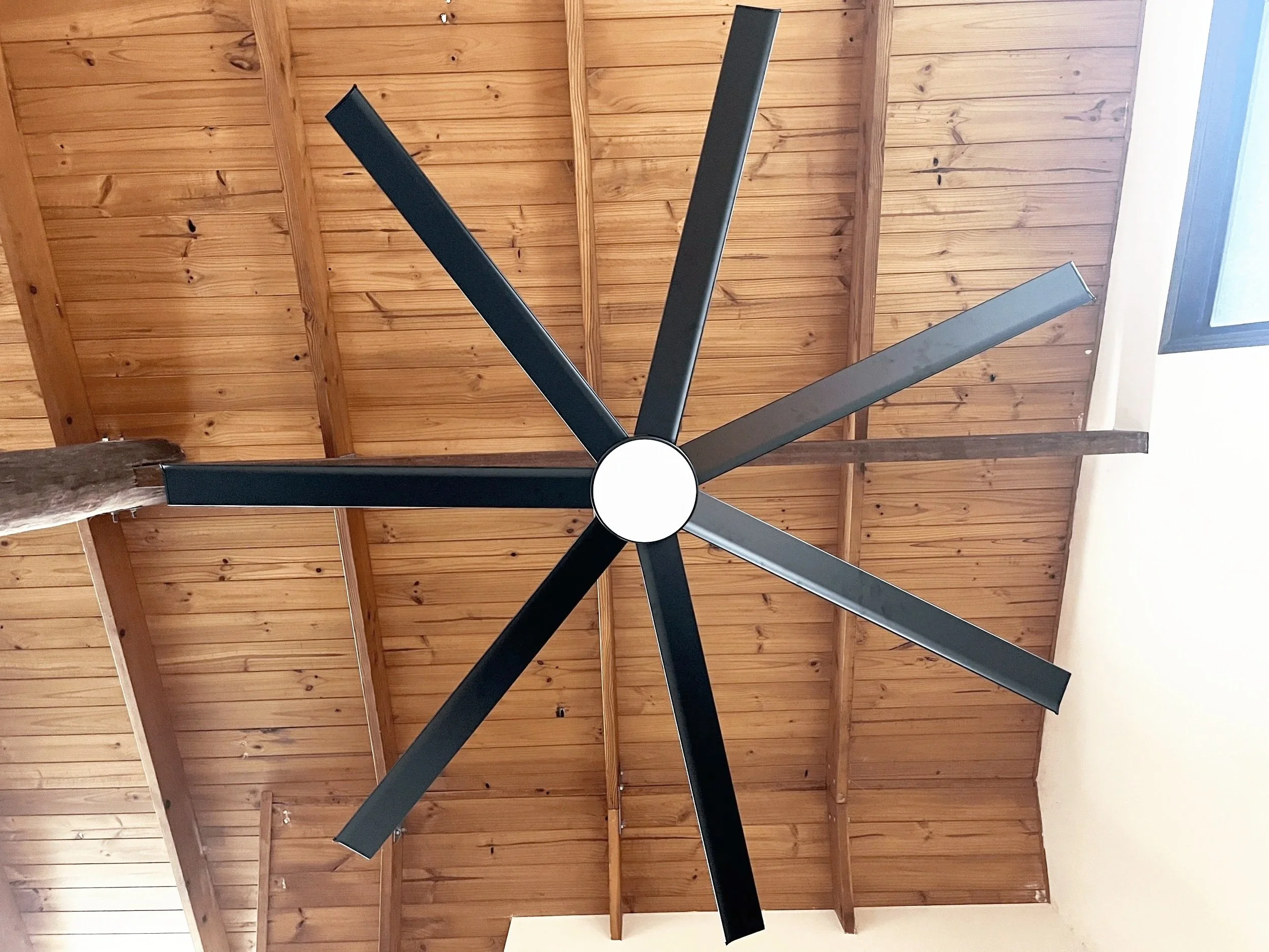 Large modern black ceiling fan with eight blades installed in a wooden ceiling with a small window nearby.