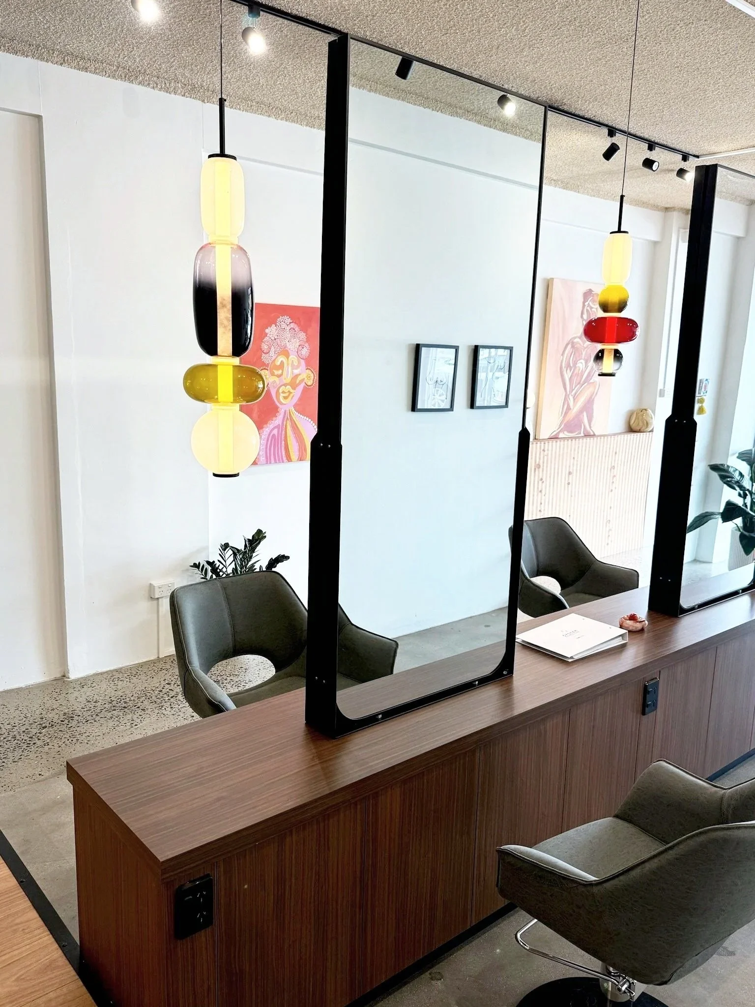 Interior of a modern salon or barbershop with three black-framed mirrors, two hanging colorful pendant lights, and a wooden counter with chairs and framed artwork on the wall.
