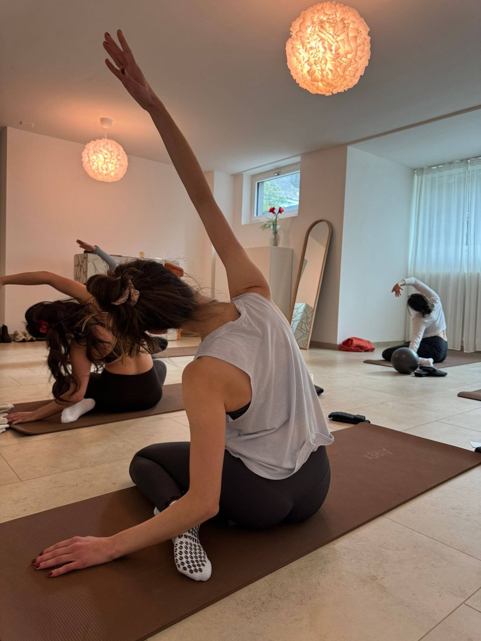 Drei Frauen machen Yoga in einem hellen Raum, auf Yoga-Matten sitzend, mit ausgestreckten Armen in einer Dehnpose.