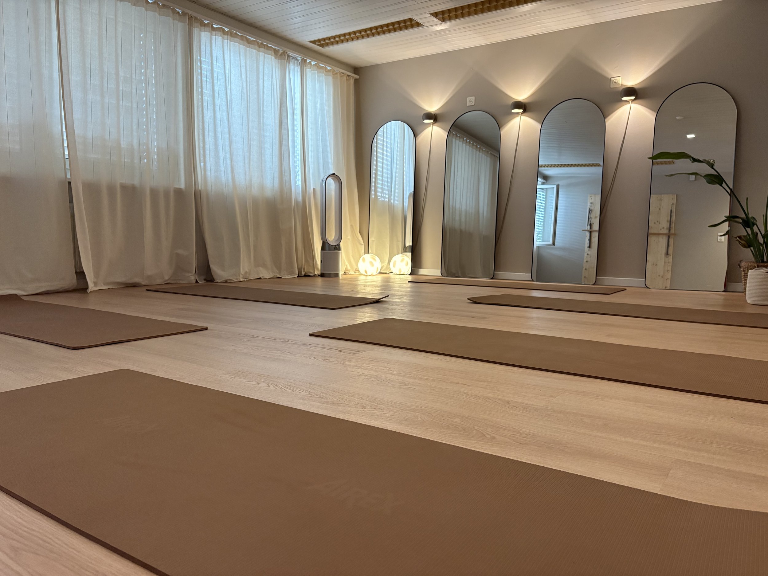 Ein Raum für Pilates, Yoga, Barre, Mobility, Wallboards, Springboards und Meditationen mit beigen Vorhängen, Spiegeln, einer Pflanze und Yoga-Matten auf dem Boden.