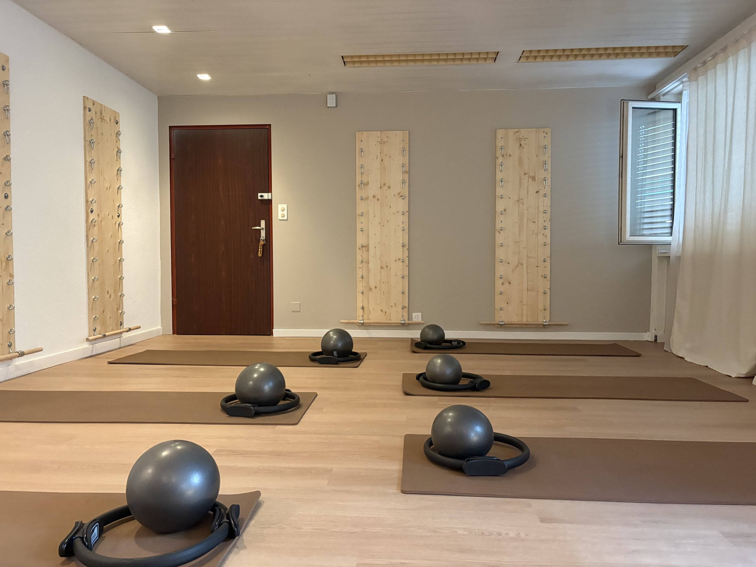 Ein Pilates- und Yogastudio mit sechs schwarzen Pilatesbällen der Marke Sissel auf beigen Airex-Yoga-Matten, an der Wand sind drei Wallboards/Springsboards angebracht, eine Tür und ein Fenster mit weißen Vorhängen sind sichtbar.